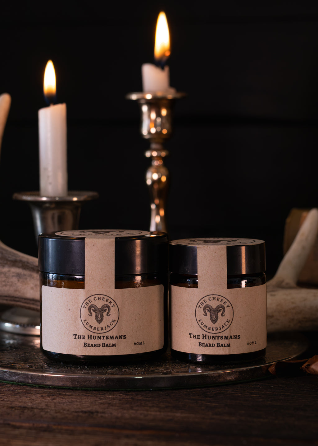 Two jars of 'The Huntsman's Beard Balm' by 'The Cheeky Lumberjack' displayed on a rustic tray with burning candles in the background.
