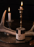 A small jar of 'The Hunter's Man' Shea Butter candle by 'Crazy Dog Candle Co.' sits on a rustic metal tray, styled with lit white candles in brass and silver holders and antlers against a dark background.