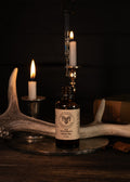 An amber glass bottle of Bear Creek Herbs 'The Huntsman's' Beard  Oil sits on a rustic metal tray, styled with antlers, lit candles, and a glass dropper in a dark, atmospheric setting.