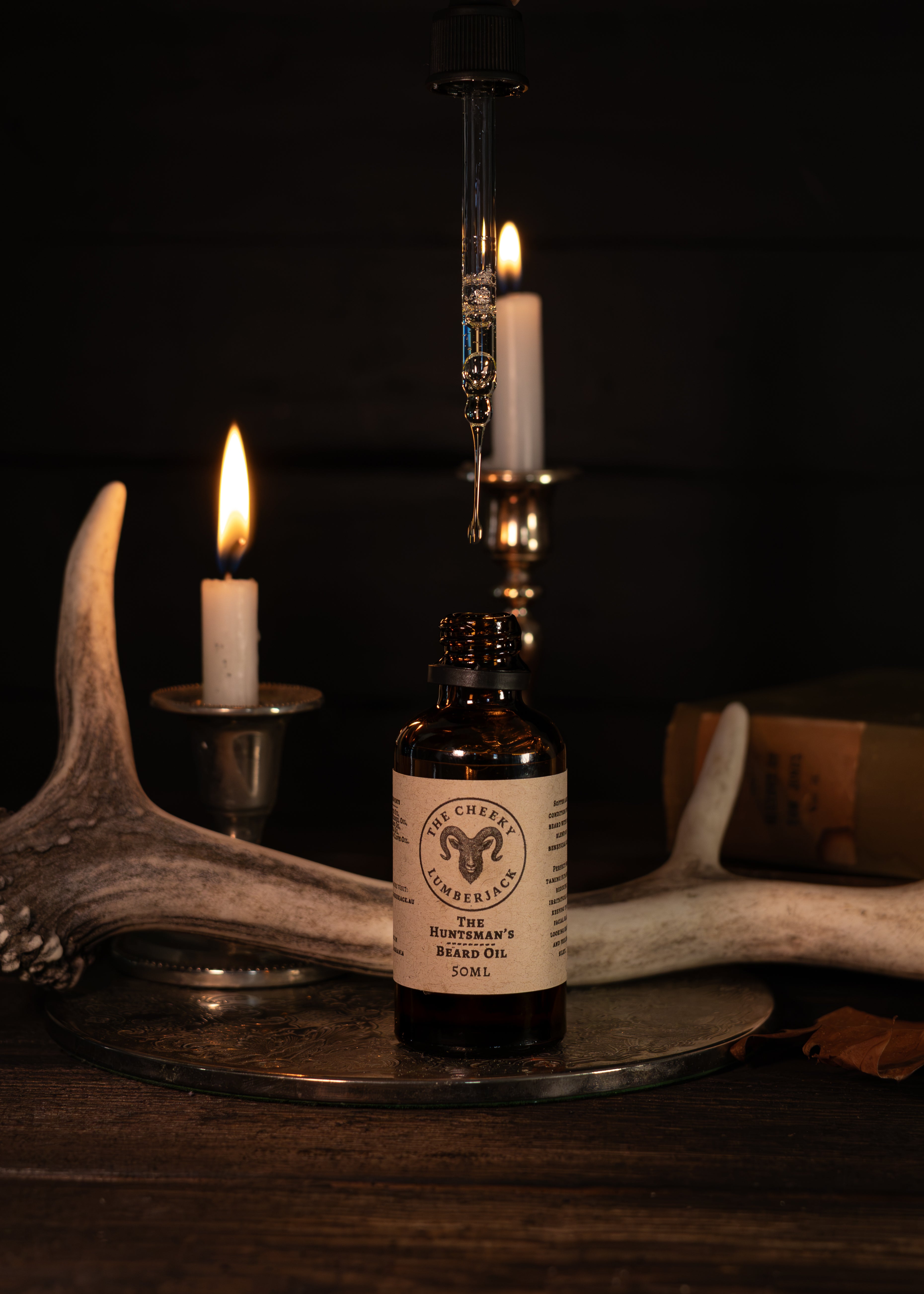 An amber glass bottle of Bear Creek Herbs 'The Huntsman's' Beard  Oil sits on a rustic metal tray, styled with antlers, lit candles, and a glass dropper in a dark, atmospheric setting.