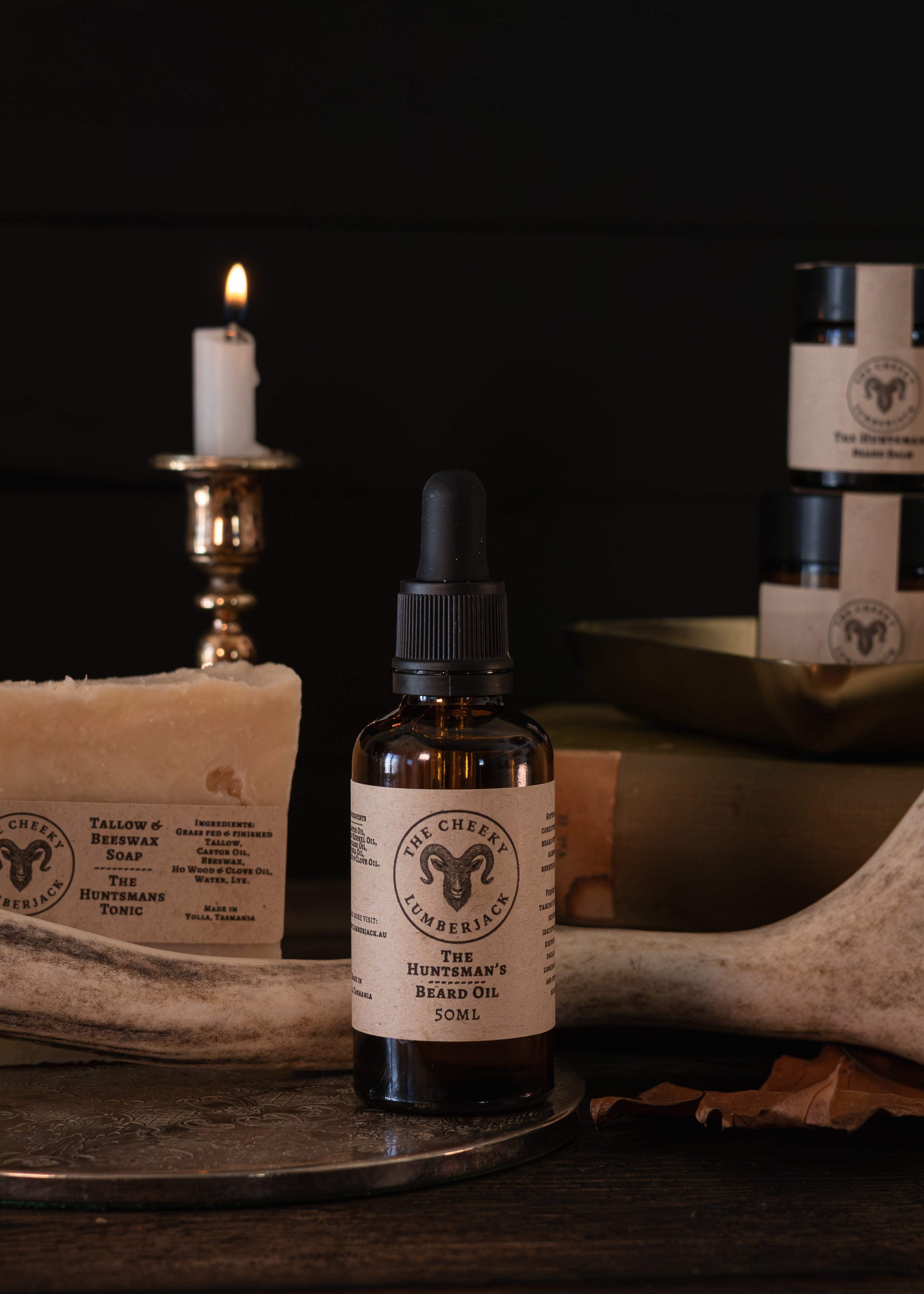 A bottle of The Huntsman's Beard Oil with a dropper cap stands on a dark wooden surface, surrounded by rustic props including a lit candle in a brass holder, a bar of soap, an antler, and other jars in the background.