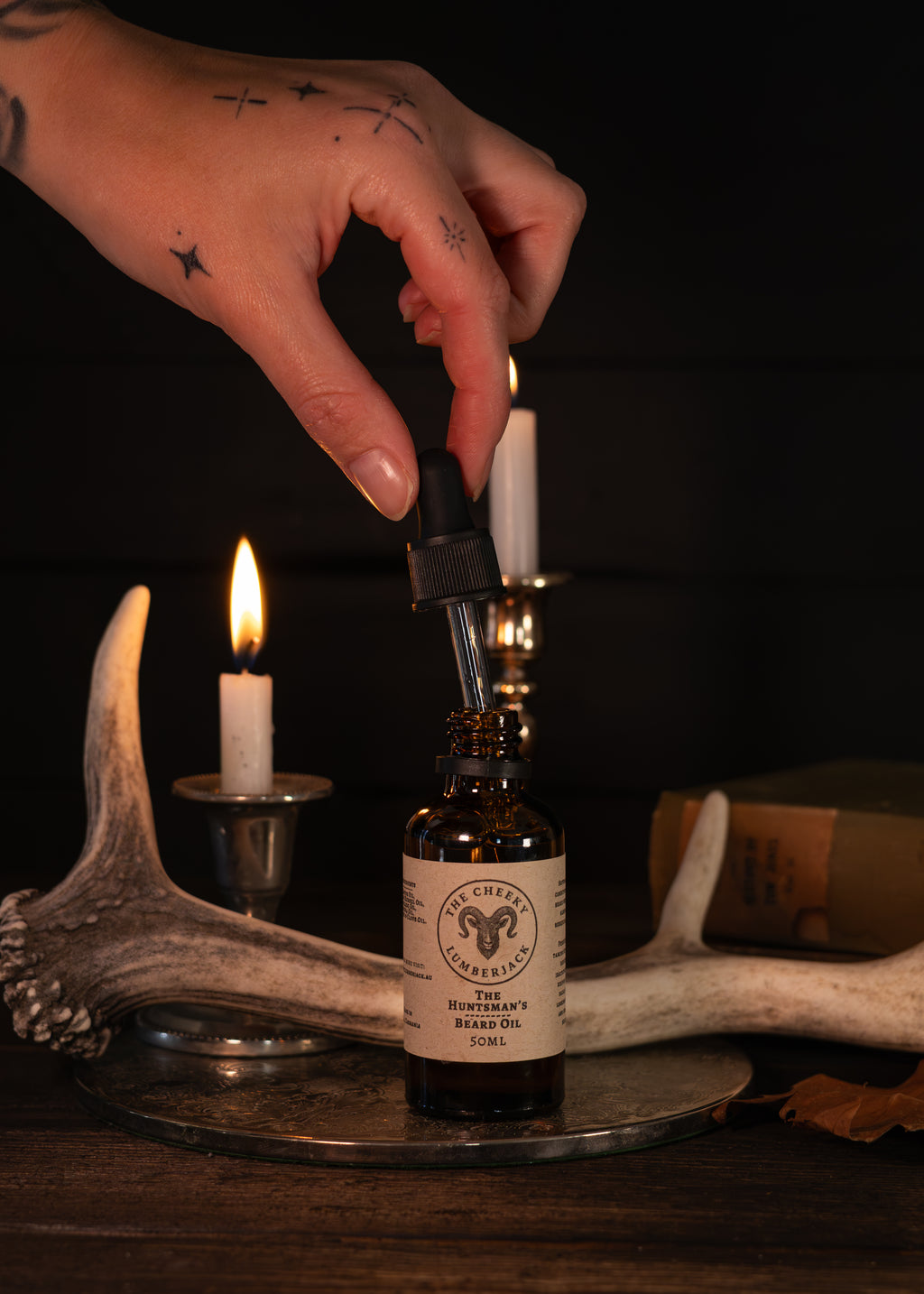A hand with tattoos holds a lit dropper above an amber glass bottle labeled 'The Cheeky Lumberjack' and 'The Huntsman's Beard Oil'. The bottle sits on a metal tray with antlers, candles, and a book in a dark rustic setting.