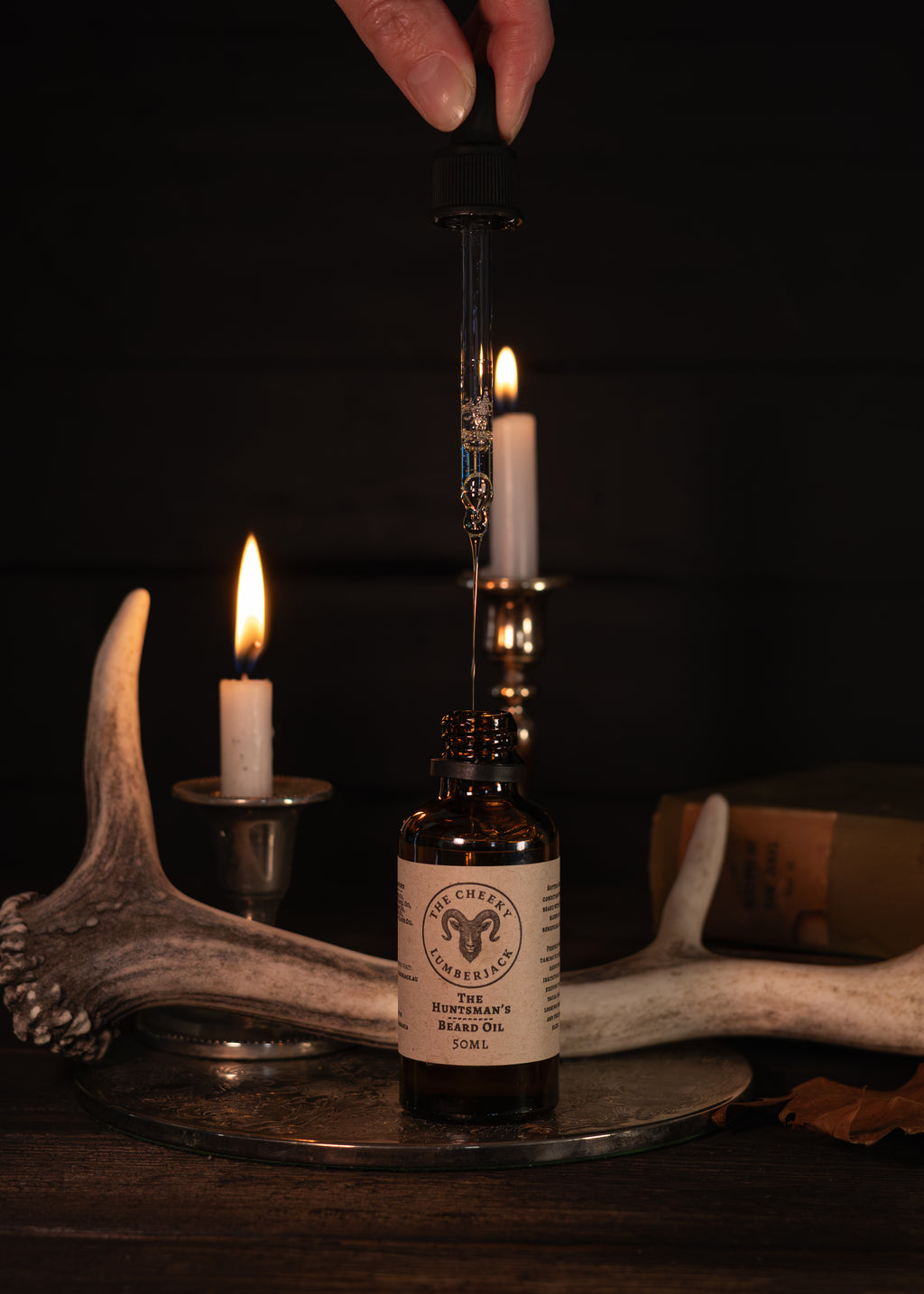 A hand holds a black dropper above an amber glass bottle labeled 'The Huntsman's Beard Oil' against a dark rustic background with lit candles, antlers, and a book.