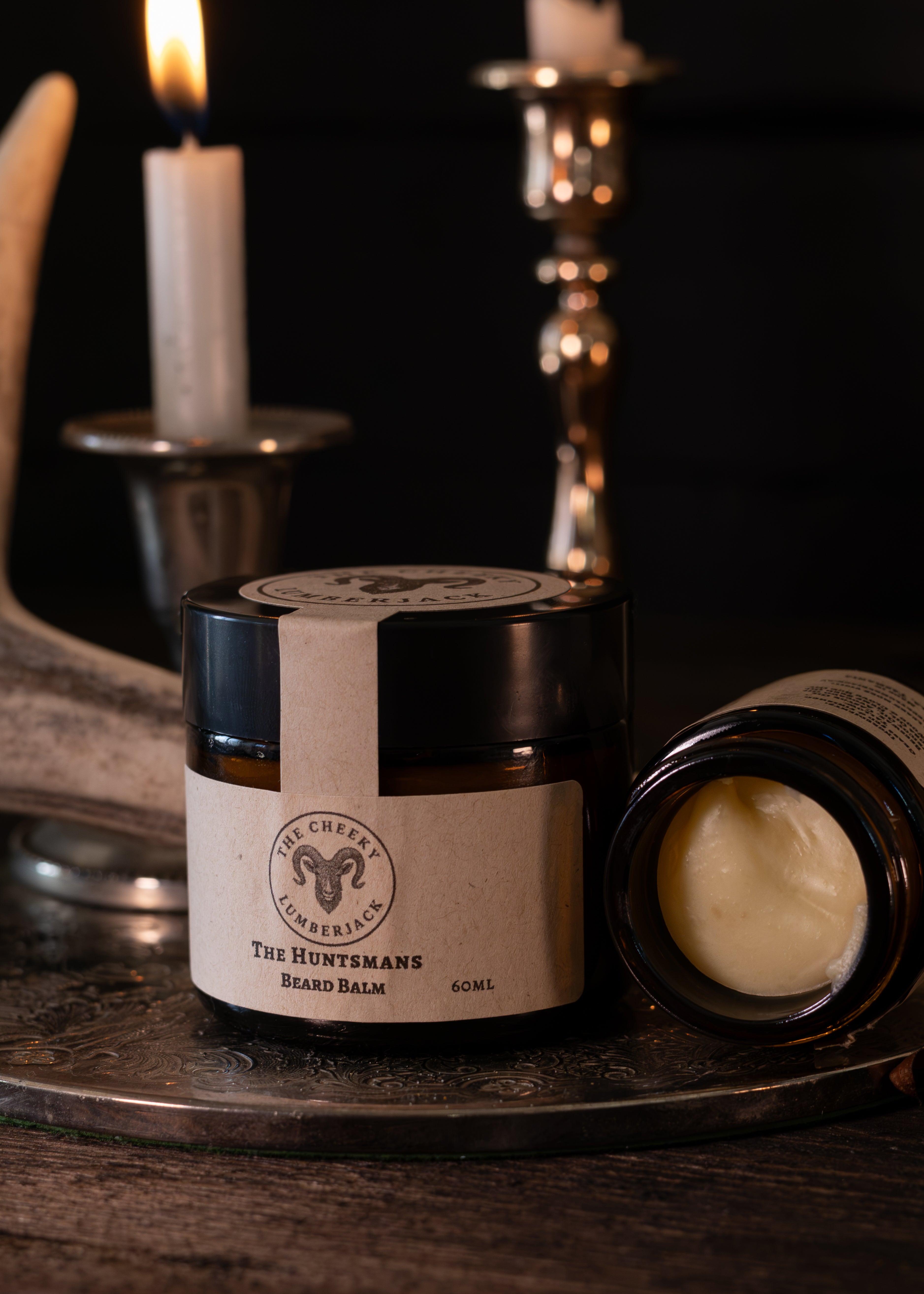 A jar of beard balm and an open jar showing the balm inside, styled on a tray with candles and metal decor in a dark, moody setting. The label reads 'The Cheeky Camper' and 'The Huntsman Beard Balm'.
