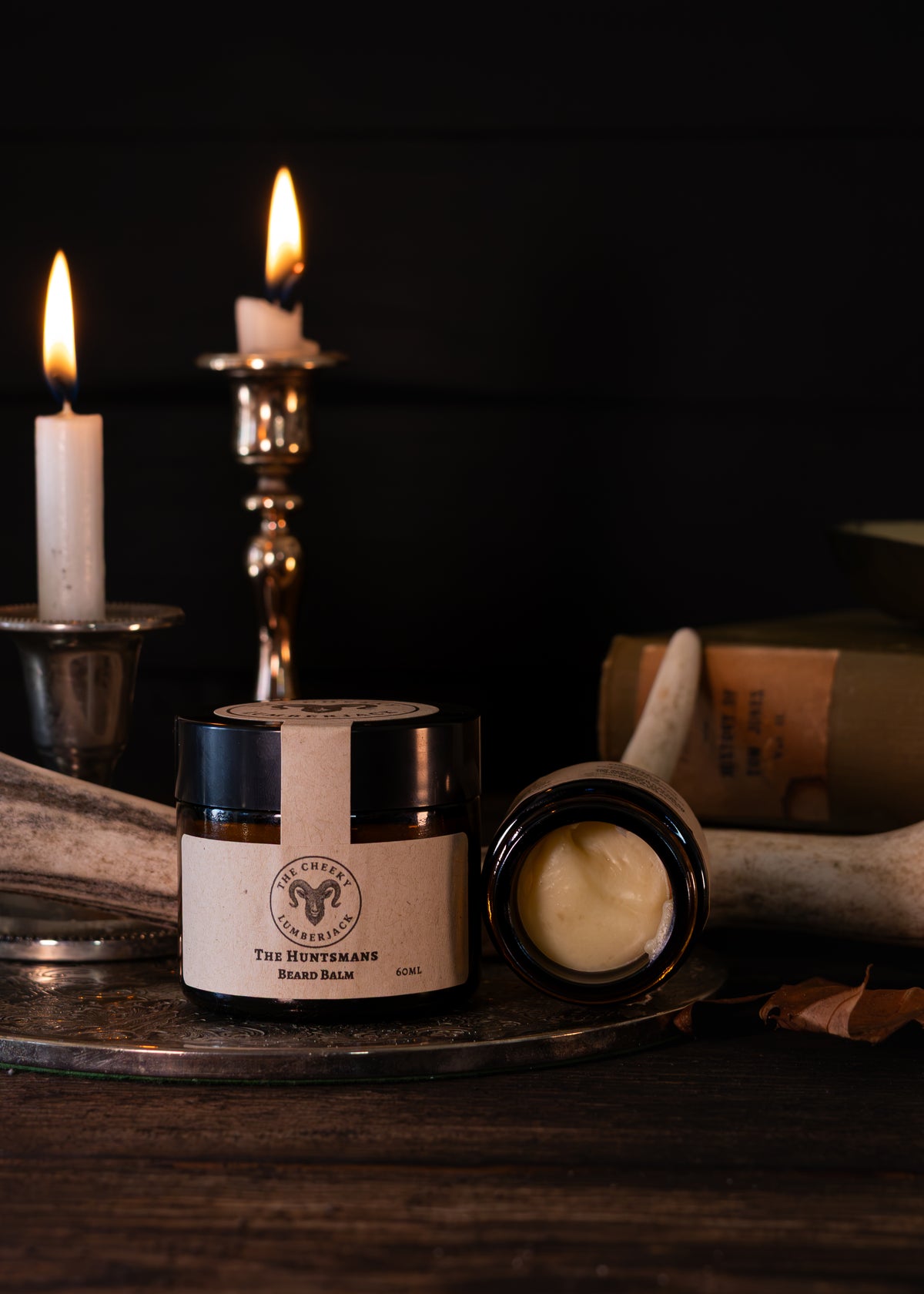 A jar of The Hunter's Beard Balm with its lid off, showing the cream inside, styled on a metal tray with lit candles, antlers, and a book in a dark, moody setting.