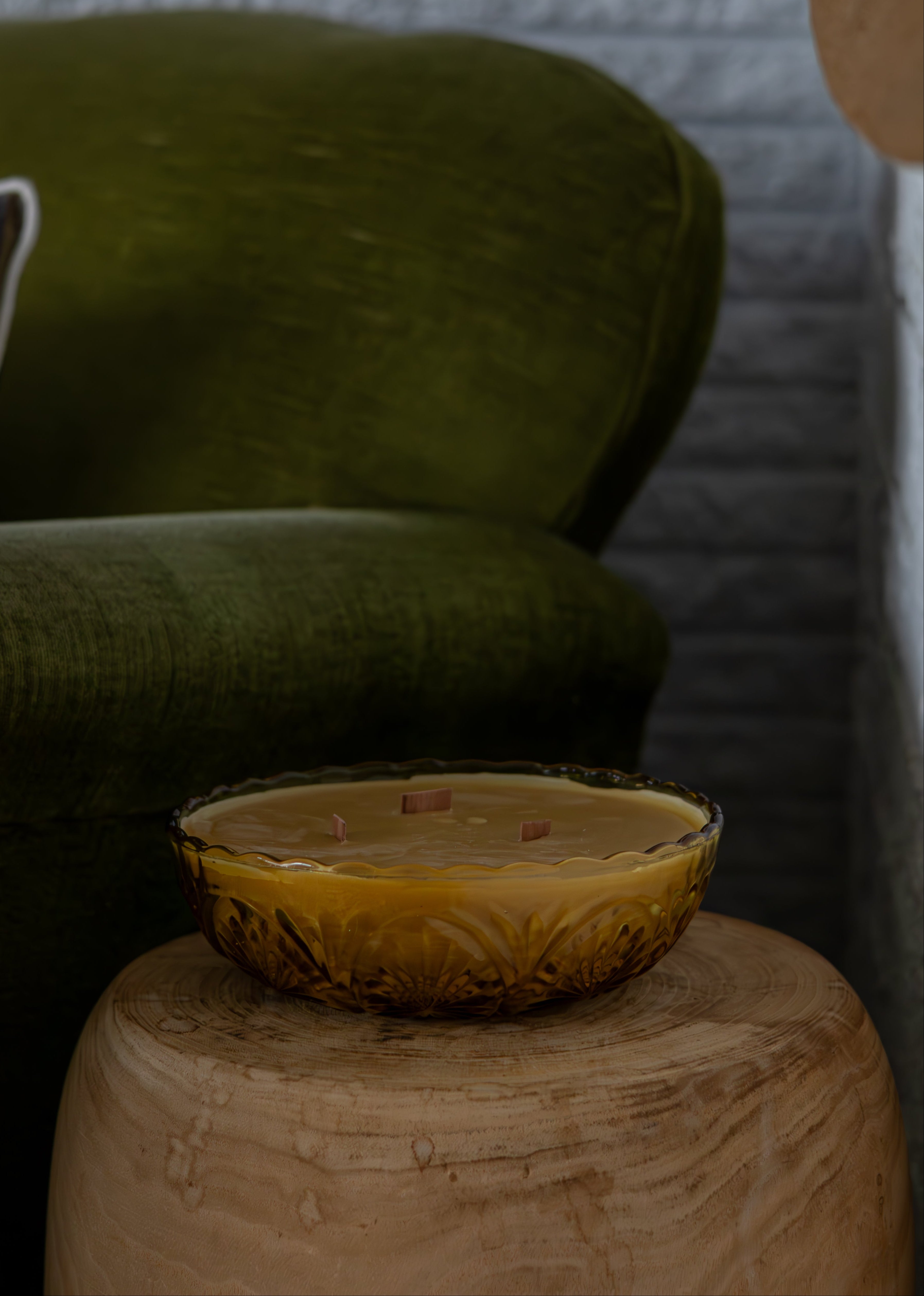 Vintage amber glass bowl beeswax candle on a wooden stool with a green cushioned chair in the background.