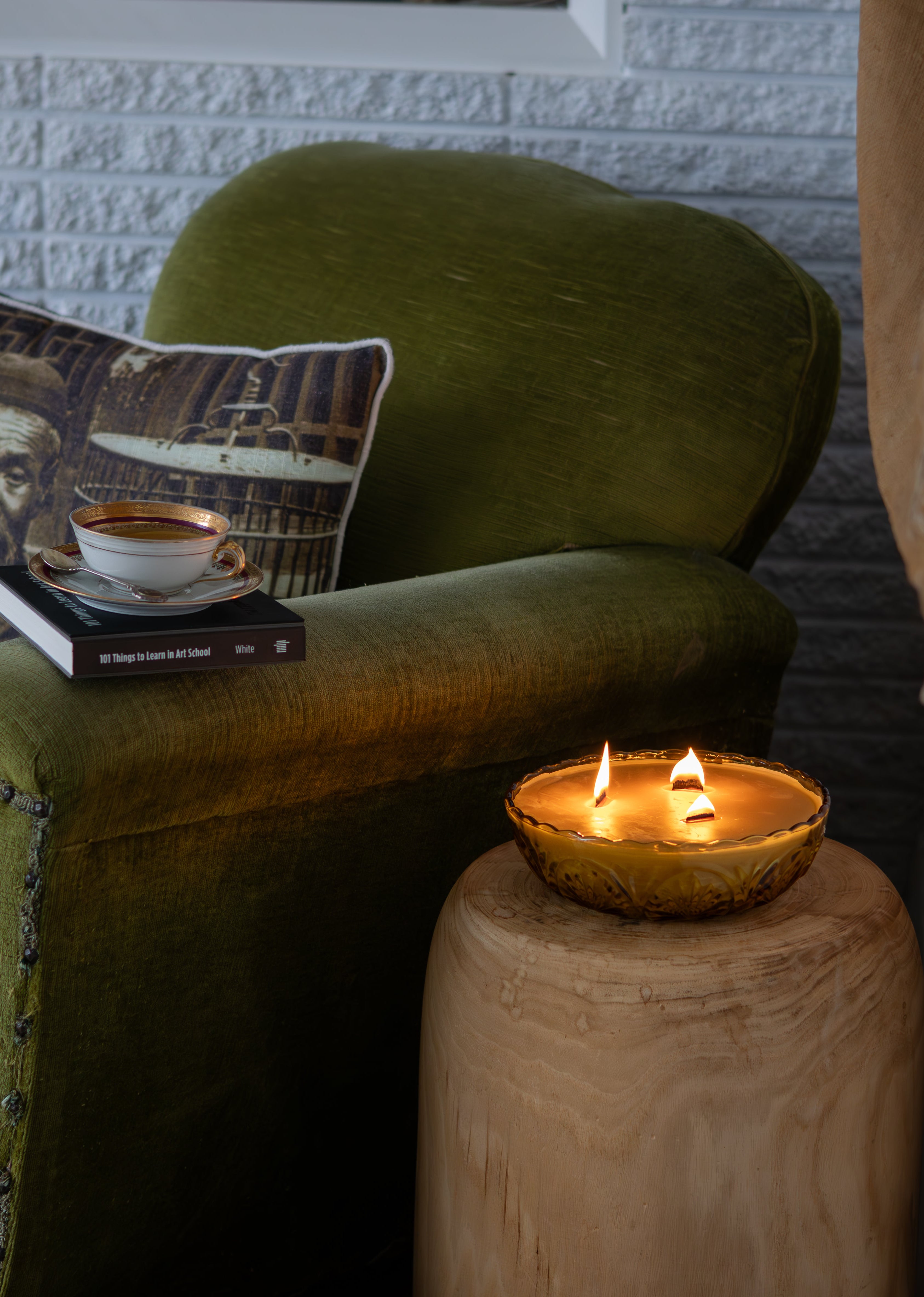 Green armchair with a pillow featuring a coffee cup, a book, and a lit vintage amber glass candle on a wooden side table.