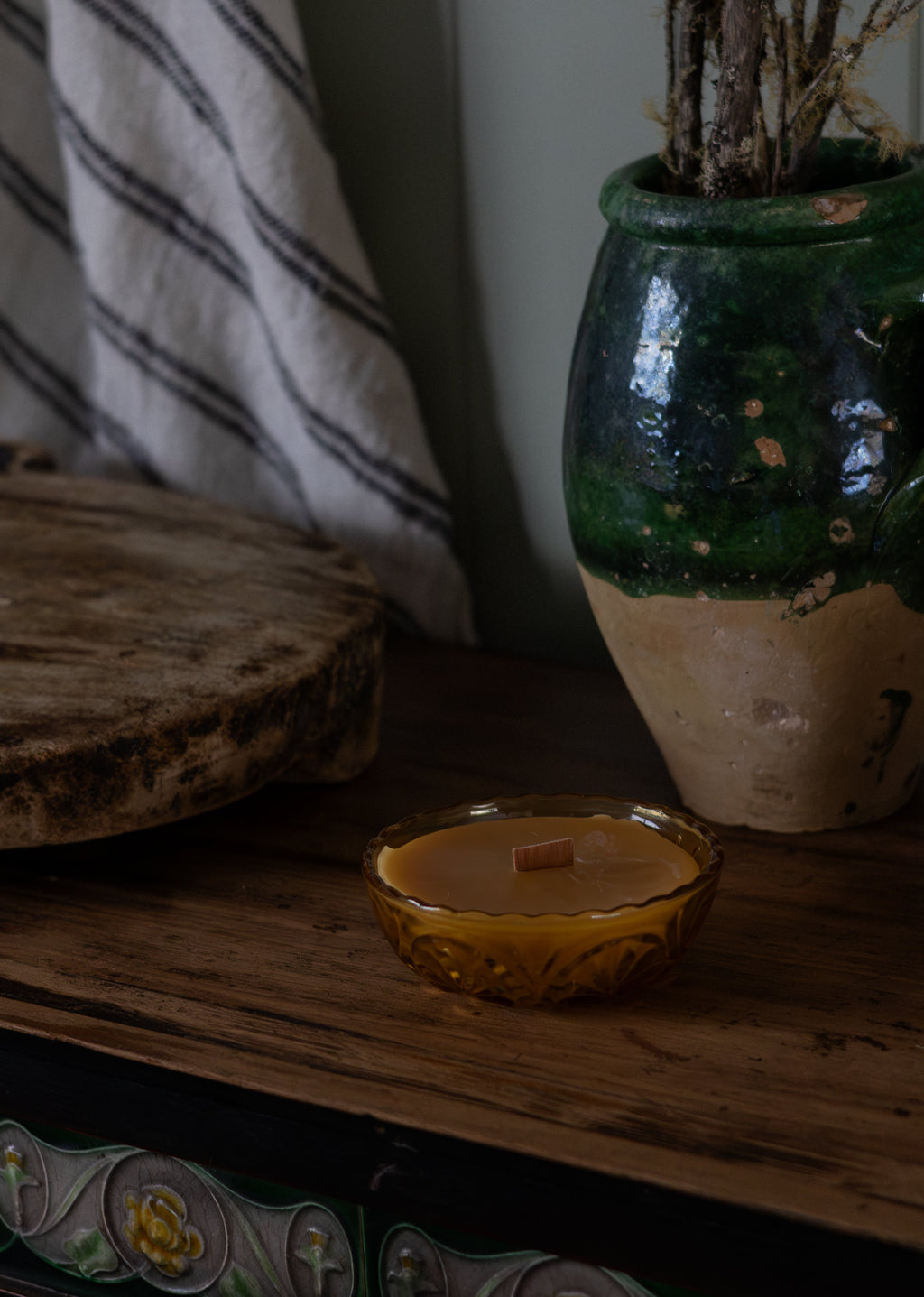 Tasmanian Beeswax Candle in a small vintage amber glass holder on a wooden surface with a decorative vase in the background.