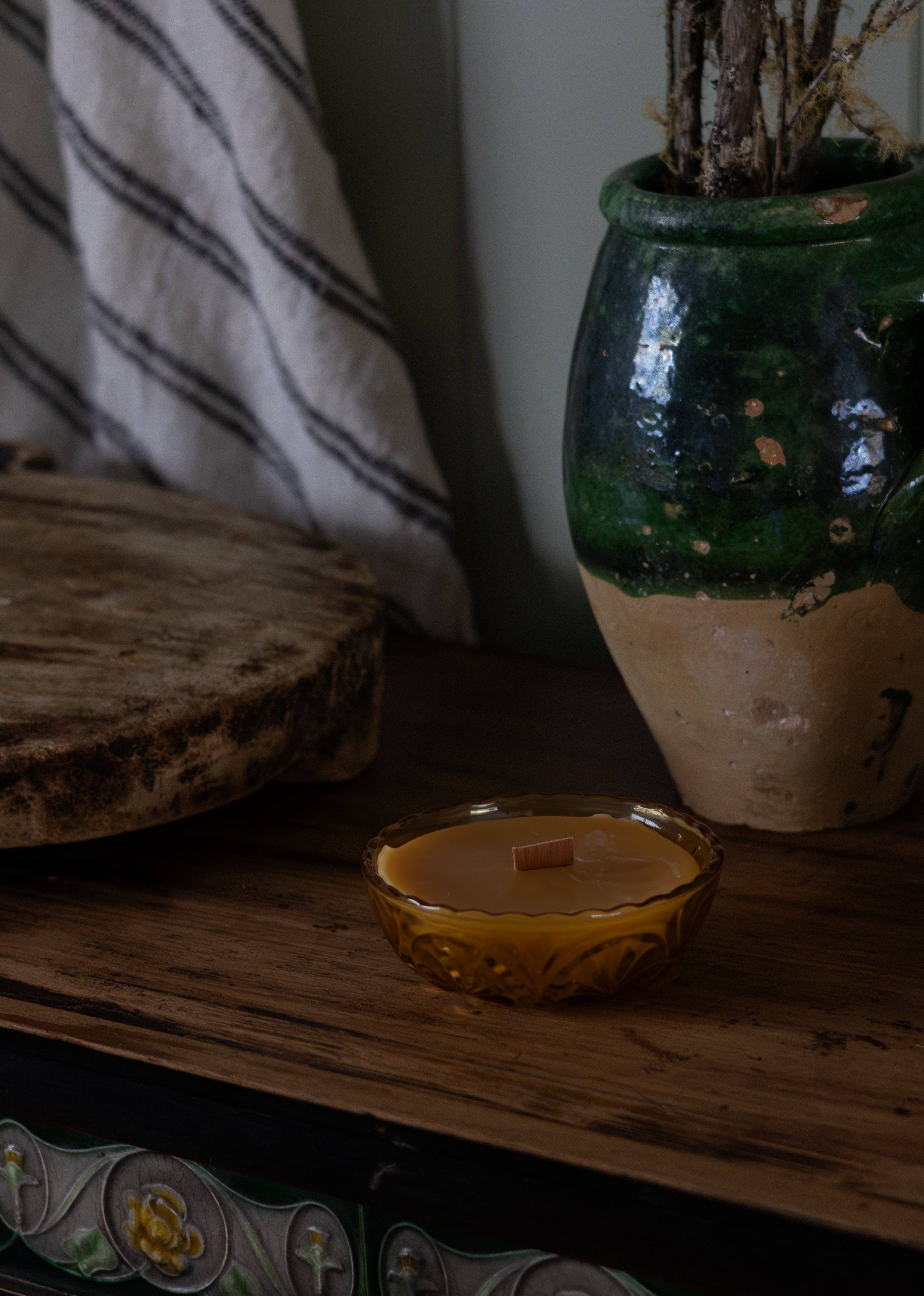 Tasmanian Beeswax Candle in a small vintage amber glass holder on a wooden surface with a decorative vase in the background.