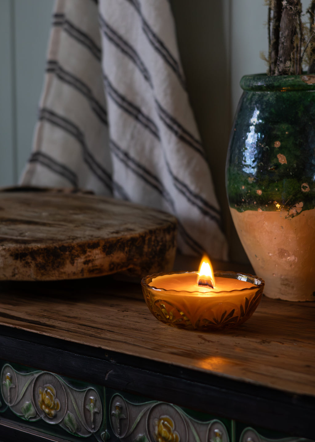Candle in a vintage amber small glass Tasmanian beeswax candle on a wooden surface with a striped cloth and decorative vase in the background.