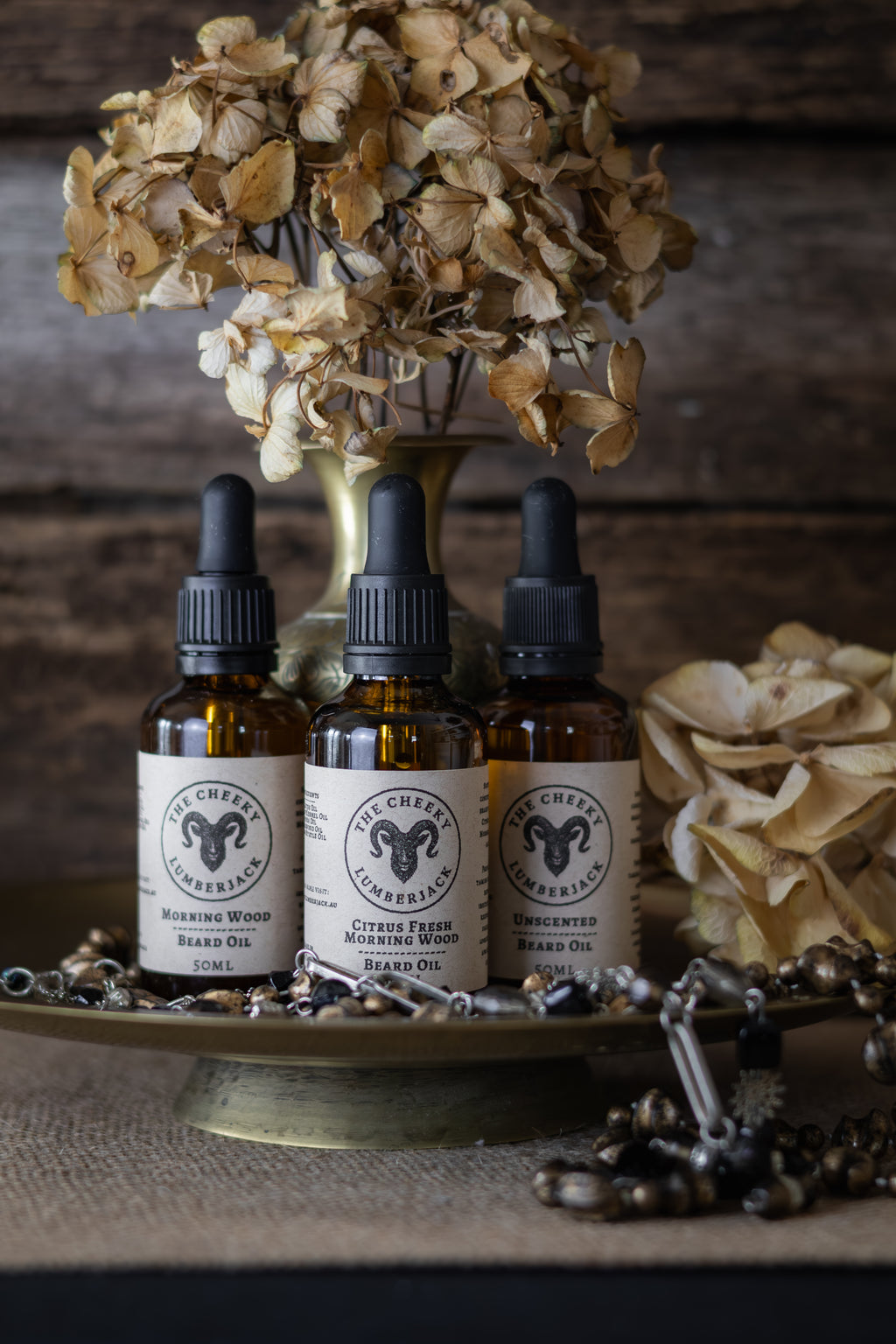 Three bottles of Cheeky Herbs brand oil on a wooden surface with dried flowers and leaves.