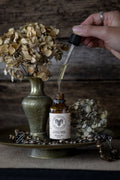 Hand using a dropper to extract oil from a bottle labeled 'Cherry & Sage' with dried flowers and a vase in the background.