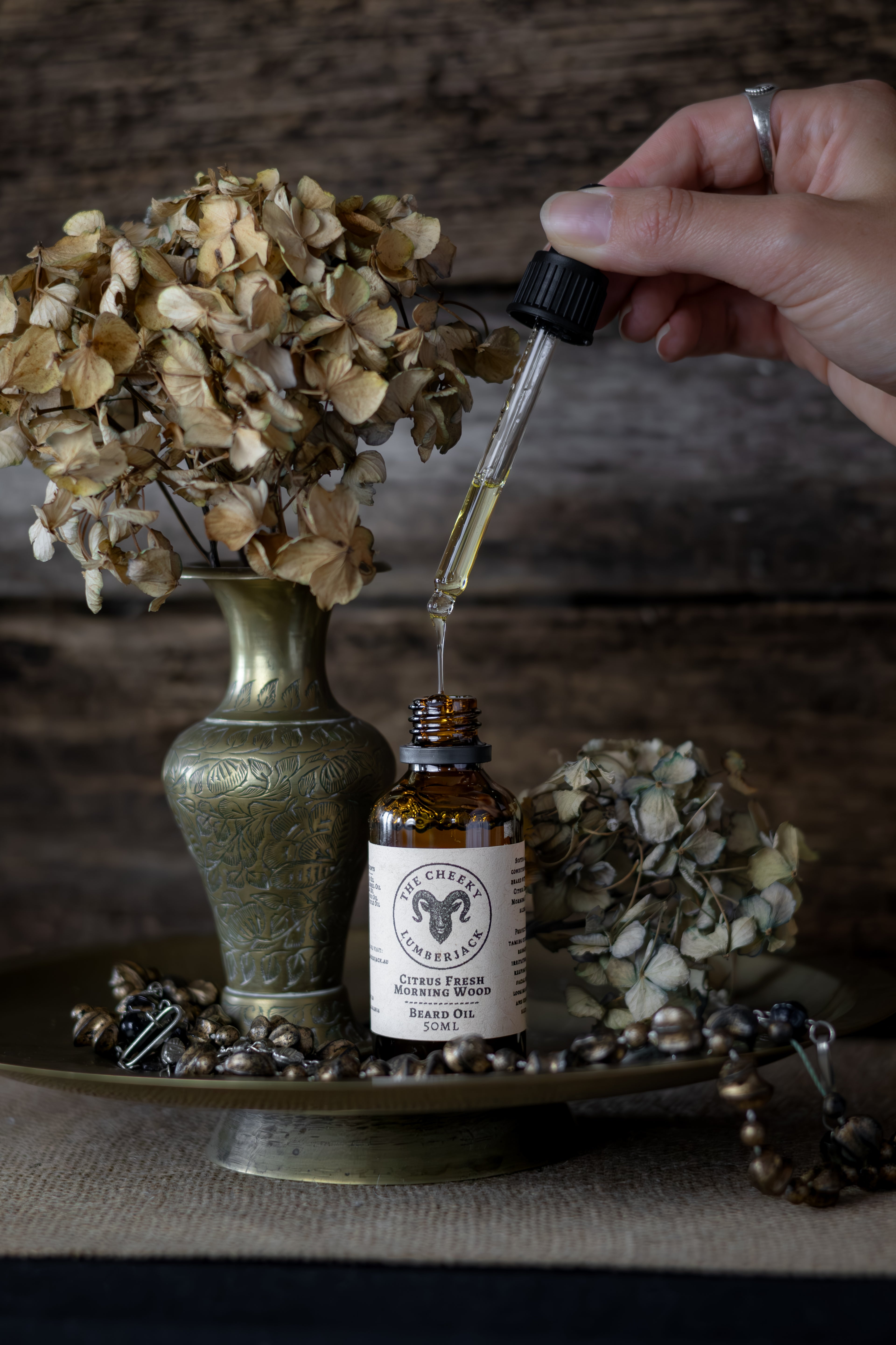 Hand using a dropper to extract oil from a bottle labeled 'Cherry & Sage' with dried flowers and a vase in the background.