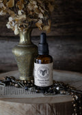 Bottle of 'Morning Wood Beard Oil' with a dropper on a rustic surface with dried flowers and jewelry.