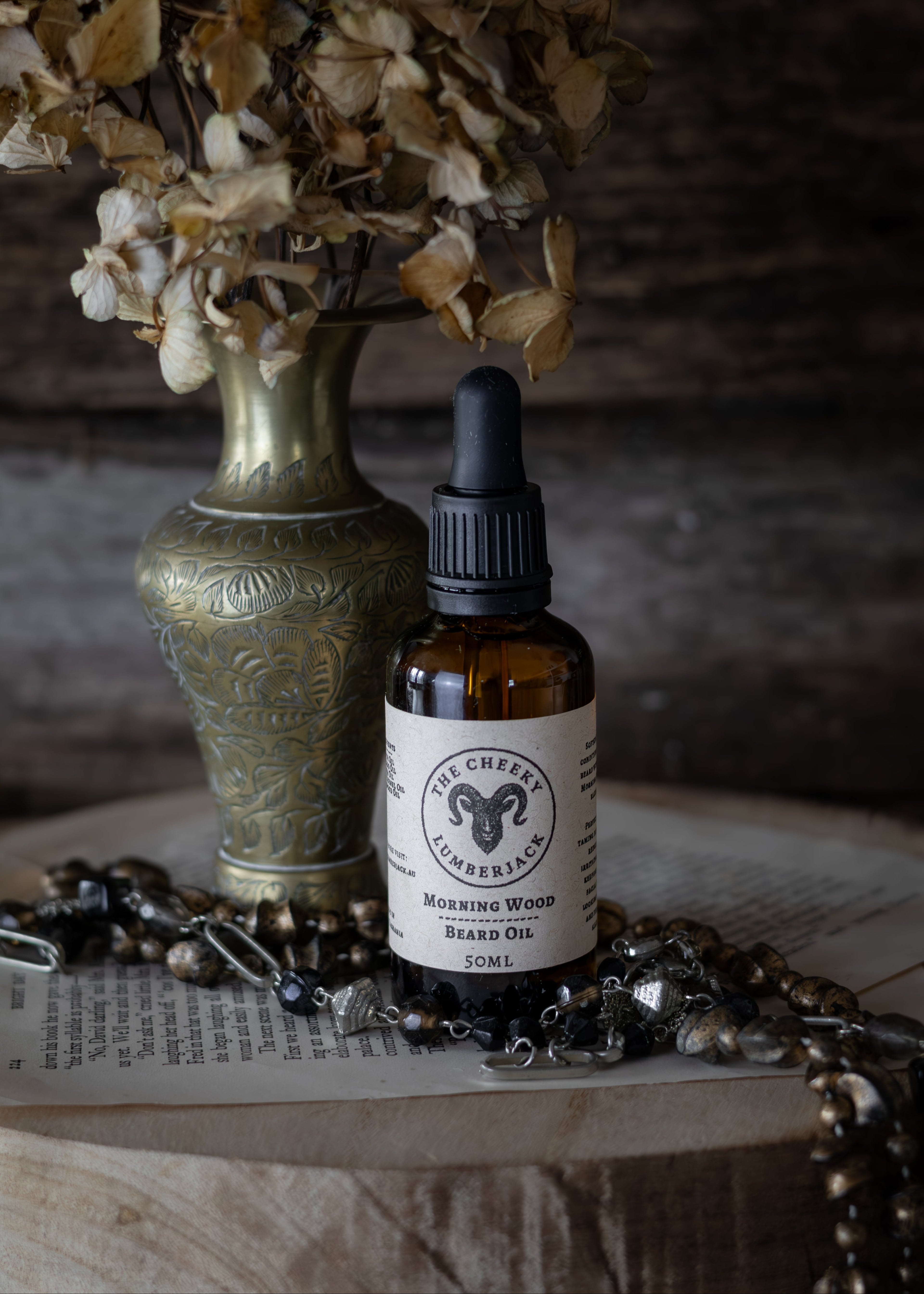 Bottle of 'Morning Wood Beard Oil' with a dropper on a rustic surface with dried flowers and jewelry.