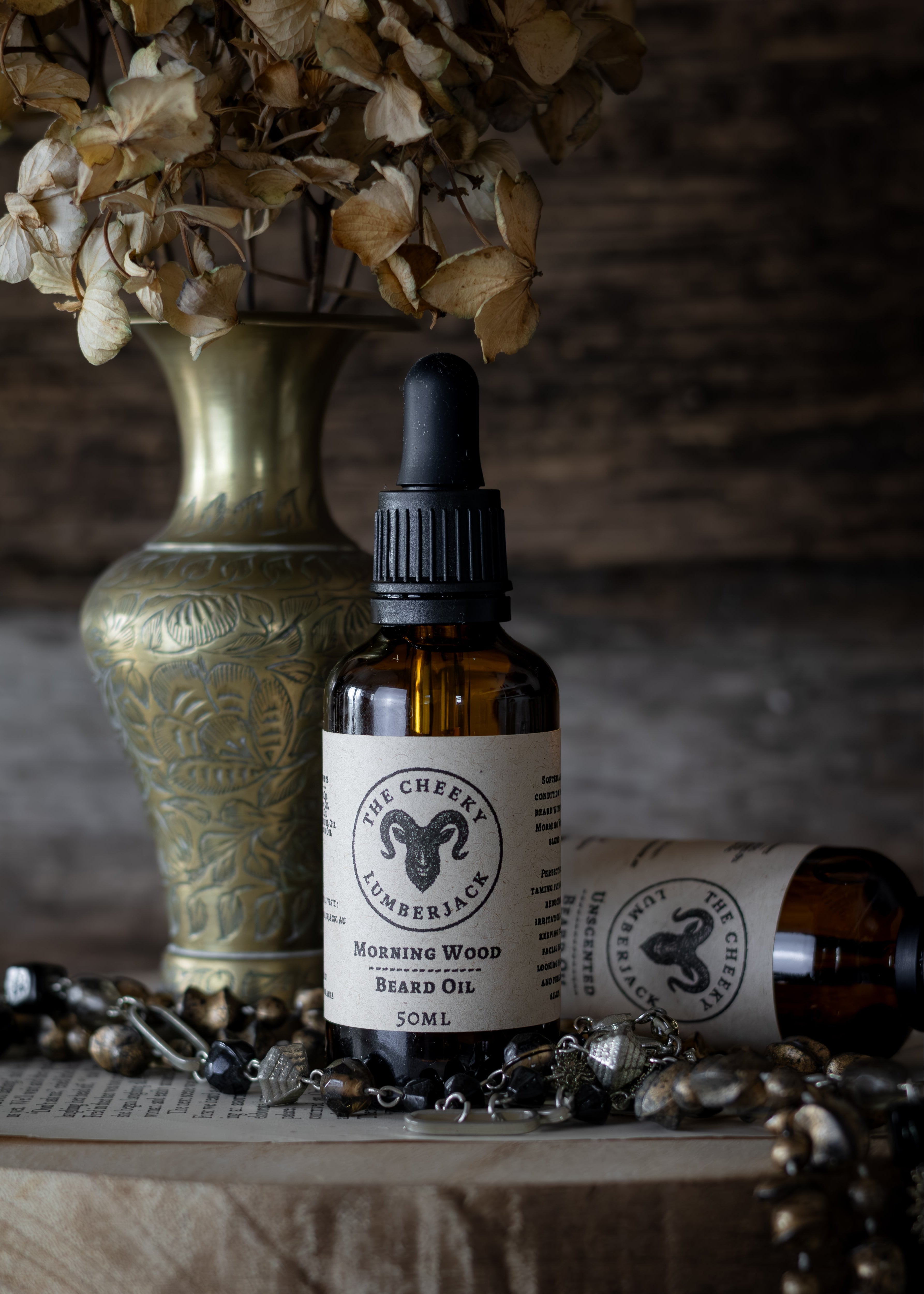 Bottle of 'Morning Wood Beard Oil' with a dropper next to a decorative vase and jewelry on a wooden surface.