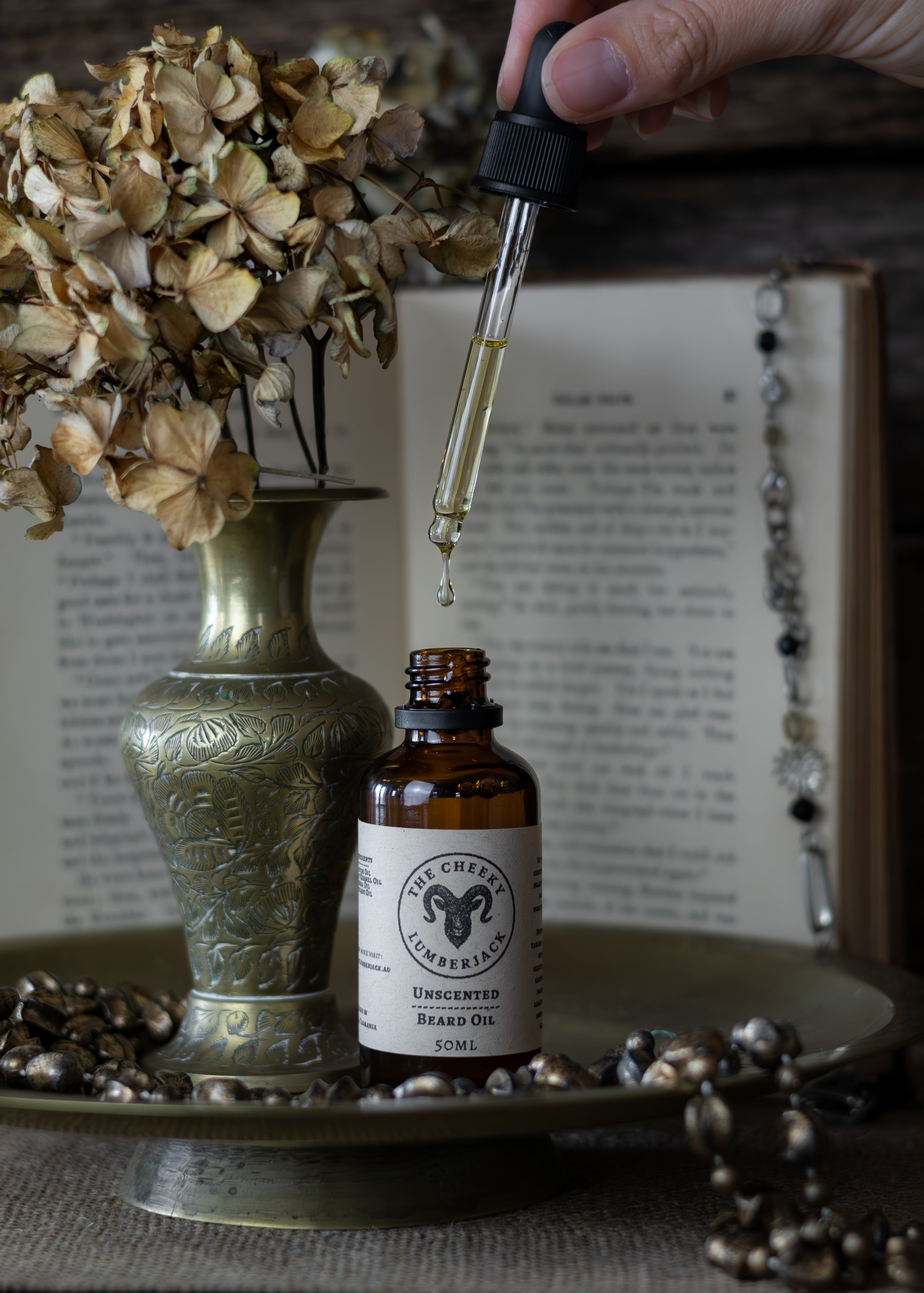 Bottle of unscented beard oil with dropper over a vase and dried flowers on a textured surface.