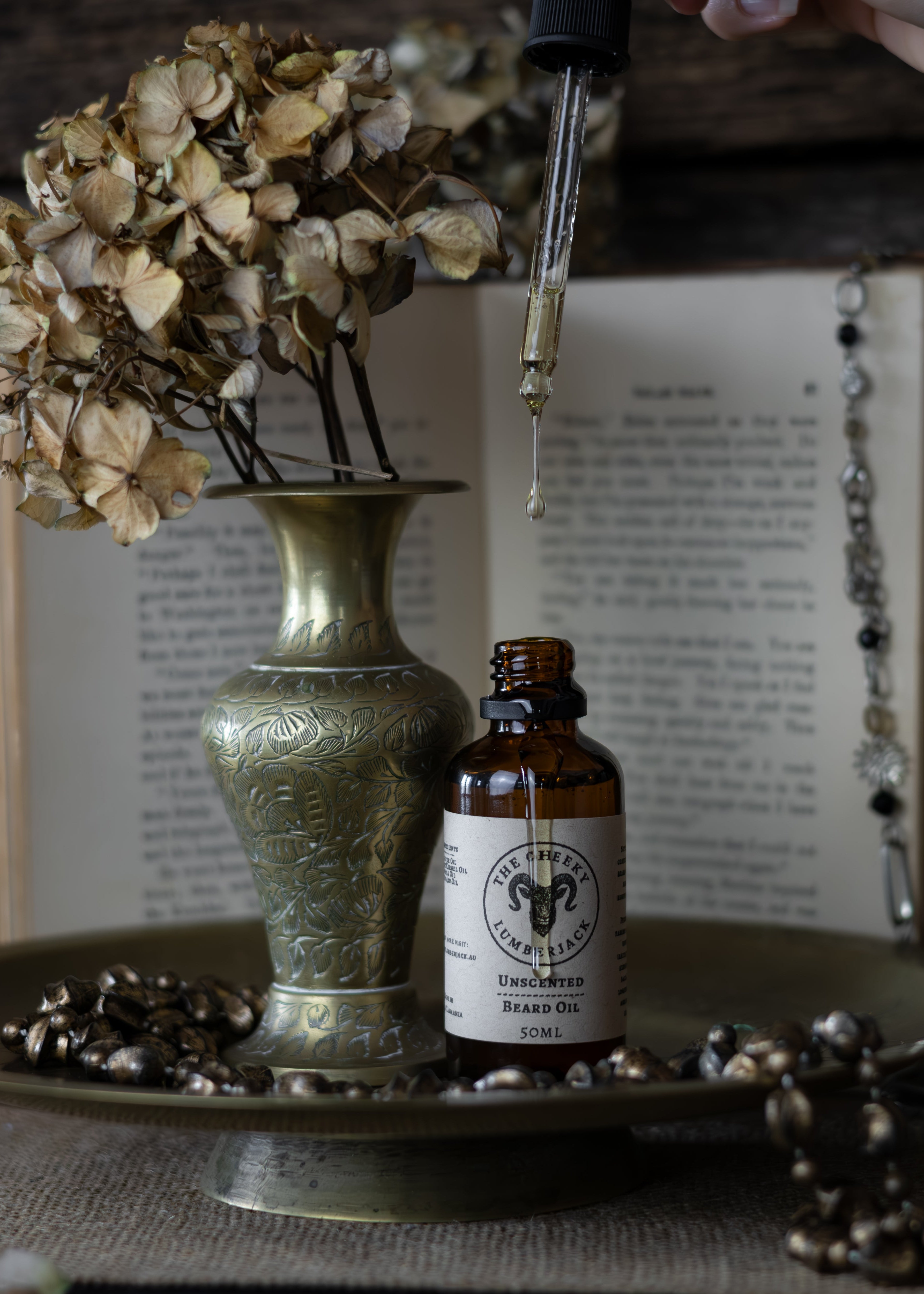 Small bottle of Unshaven brand oil with dropper on a tray with dried flowers and an open book in the background.