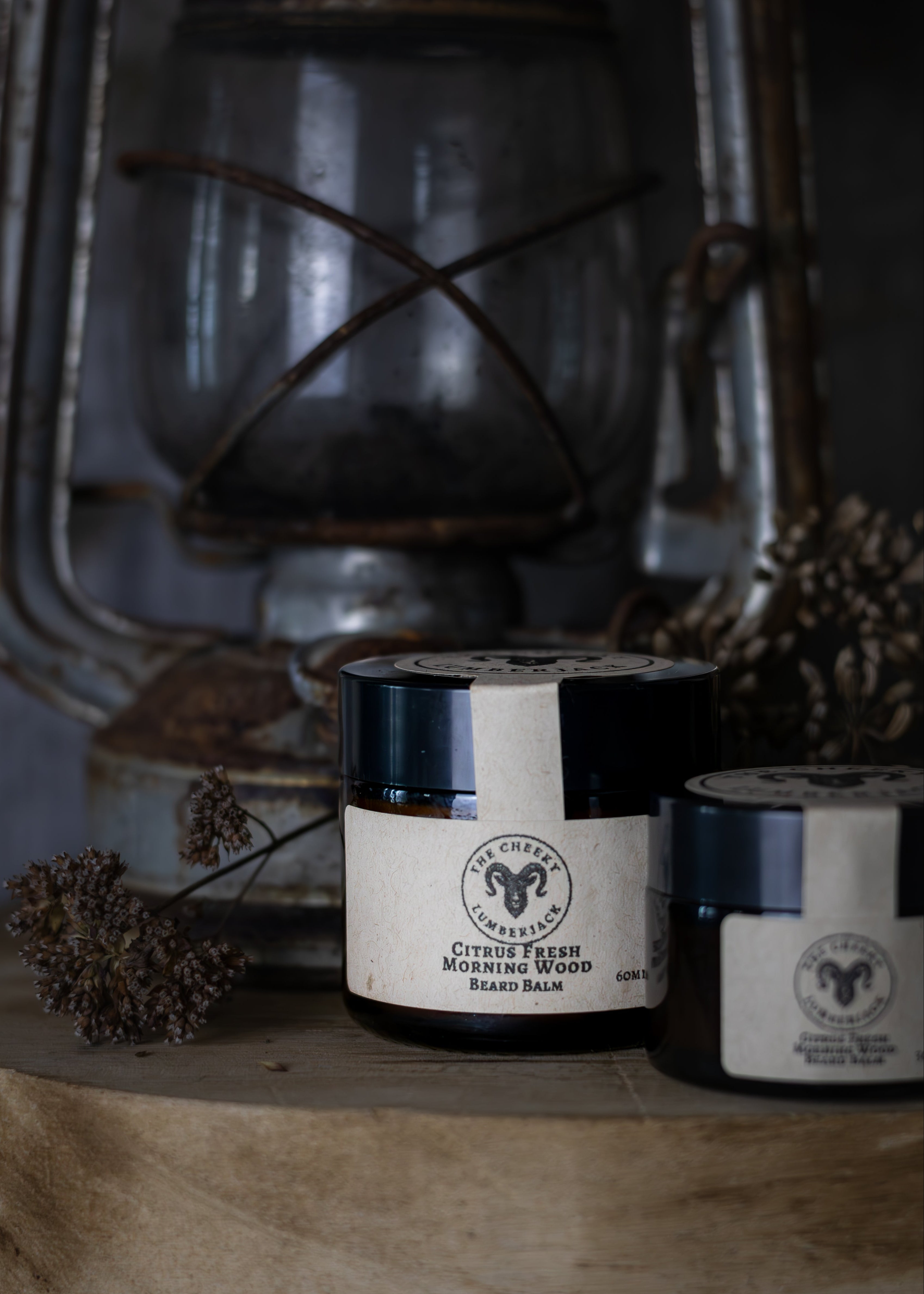 Two containers of beard balm on a wooden surface with a lantern in the background
