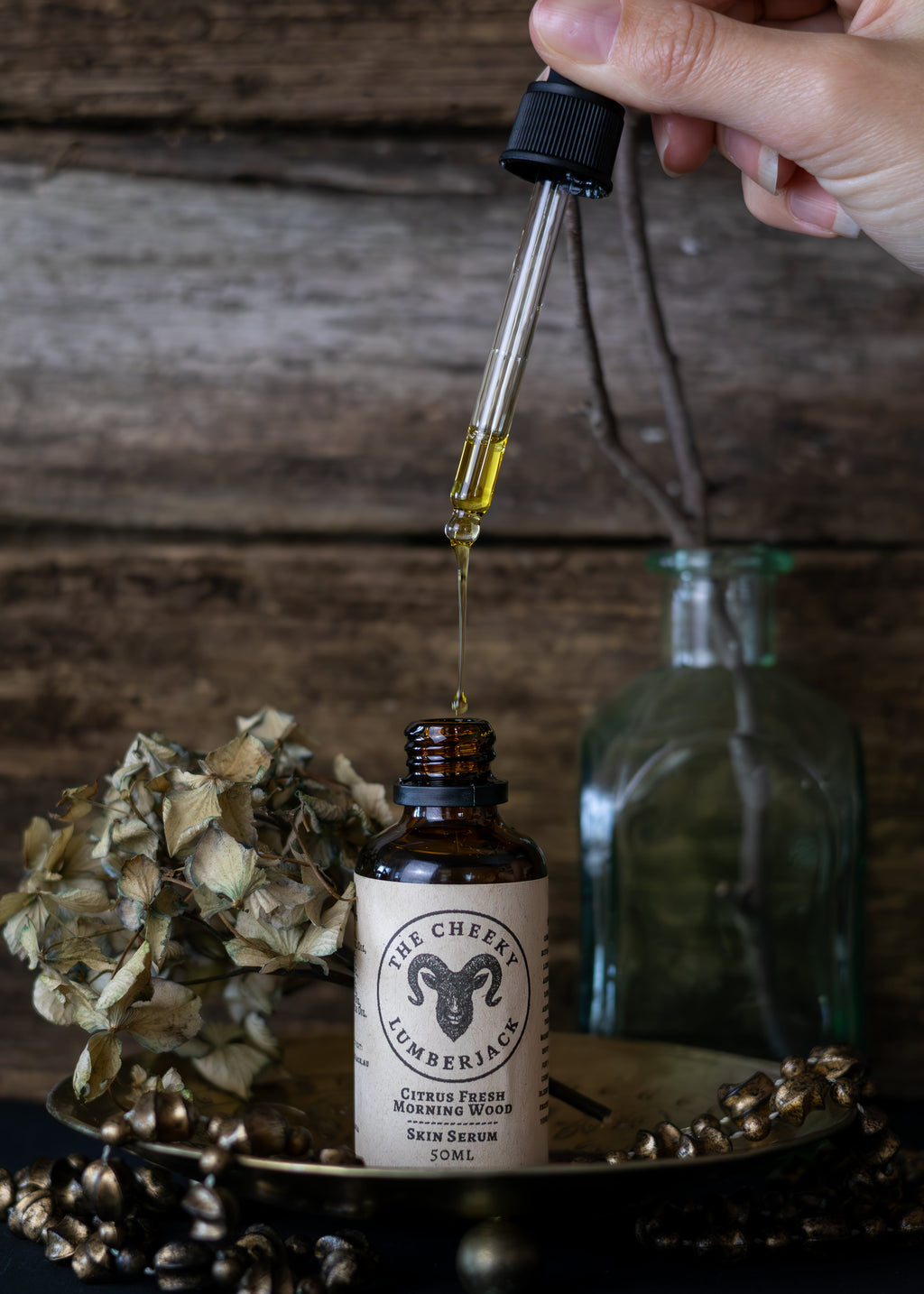 Hand holding a dropper over a bottle labeled 'The Cheeky Lumberjack' with a rustic background.