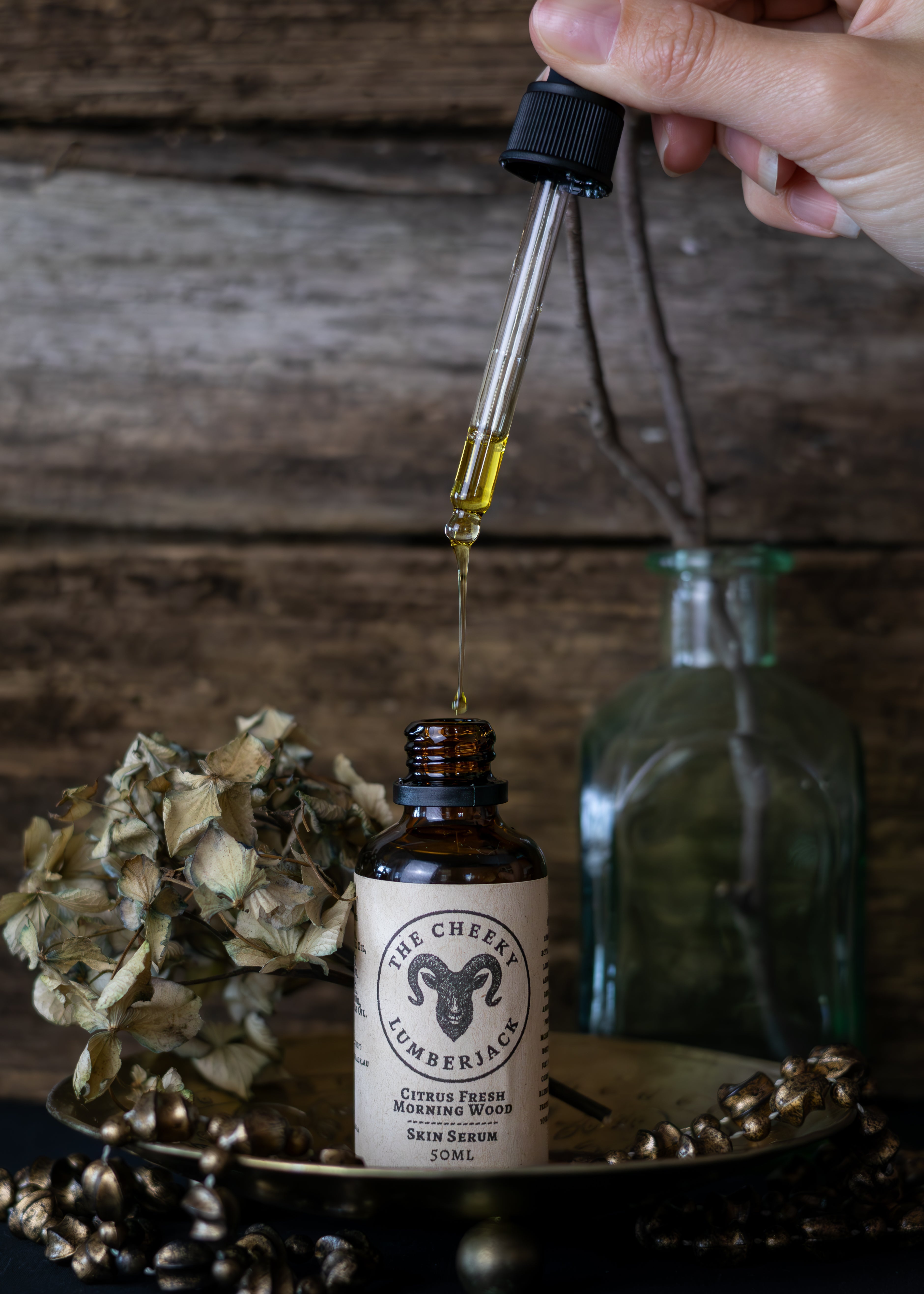 Hand holding a dropper over a bottle labeled 'The Cheeky Lumberjack' with a rustic background.