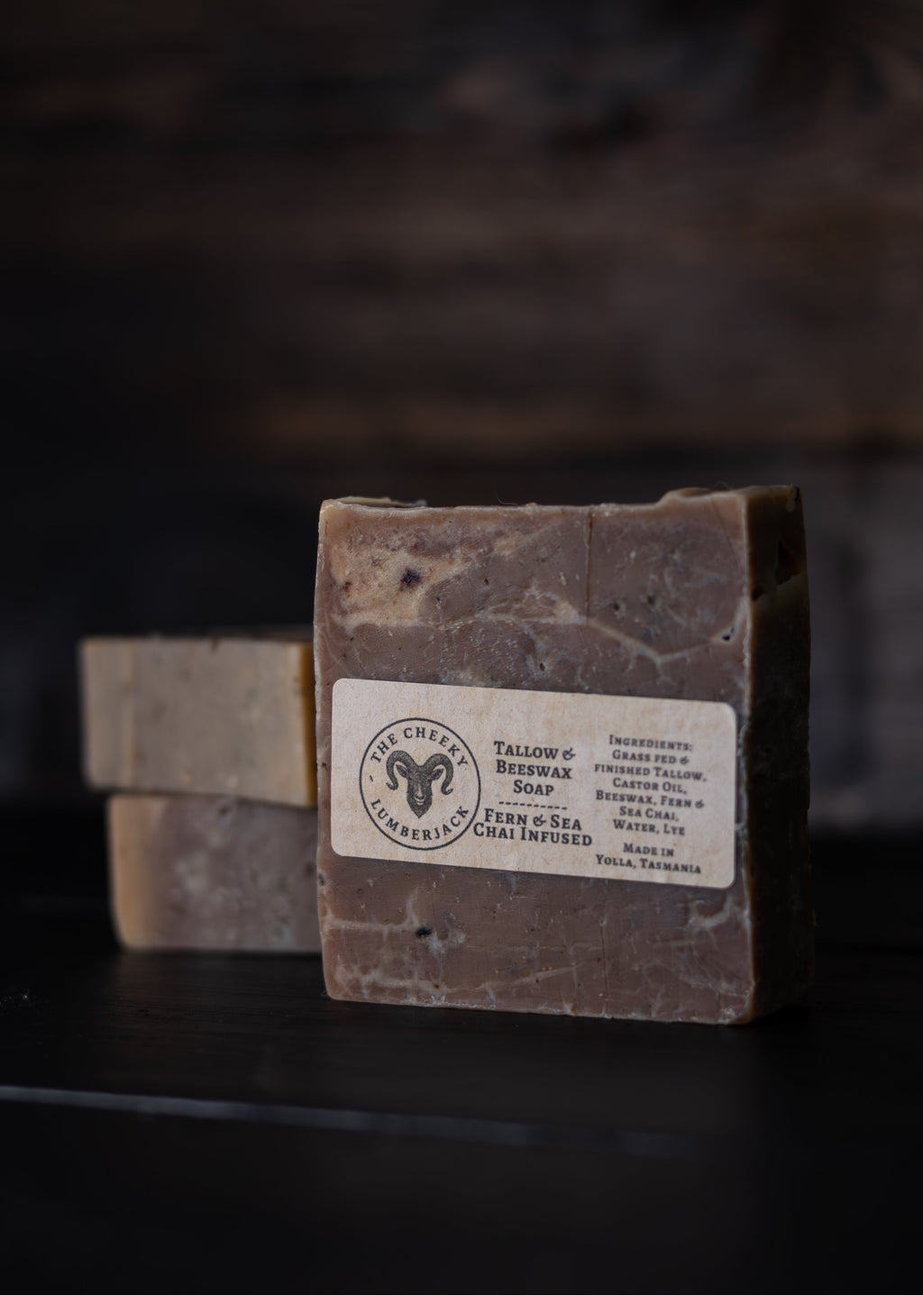 A rectangular bar of soap standing upright on a dark surface, with two additional soap bars blurred in the background. The front bar has a white label reading 'Tallow & Sea Cold Process Soap' with a ram logo and ingredients including tallow, coconut oil, and sea salt.
