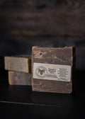A rectangular bar of soap standing upright on a dark surface, with two additional soap bars blurred in the background. The front bar has a white label reading 'The Creamy Sheep' and 'Tallow & Bee wax Soap'.