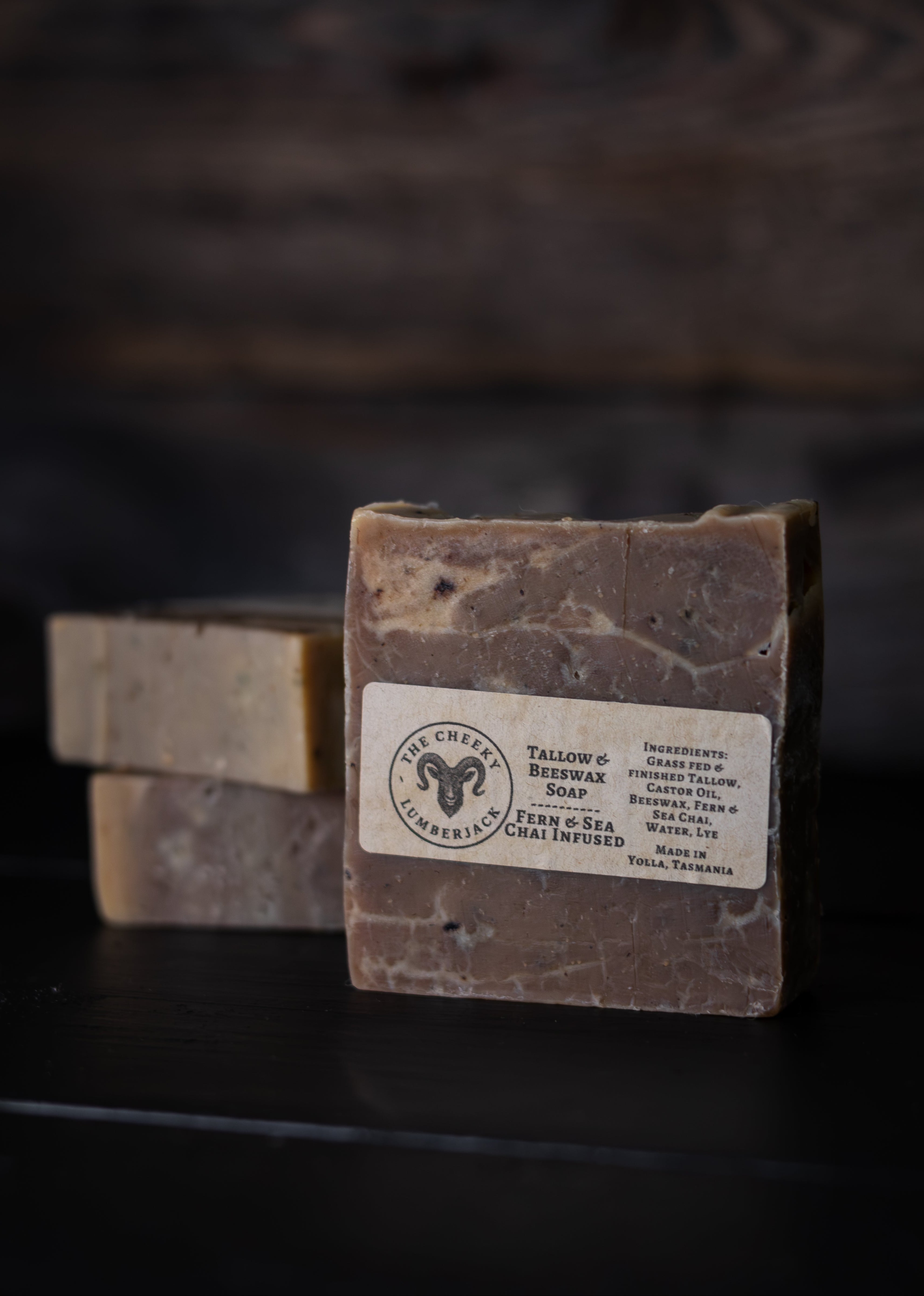 A rectangular bar of soap standing upright on a dark surface, with two additional soap bars blurred in the background. The front bar has a white label reading 'The Creamy Sheep' and 'Tallow & Bee wax Soap'.