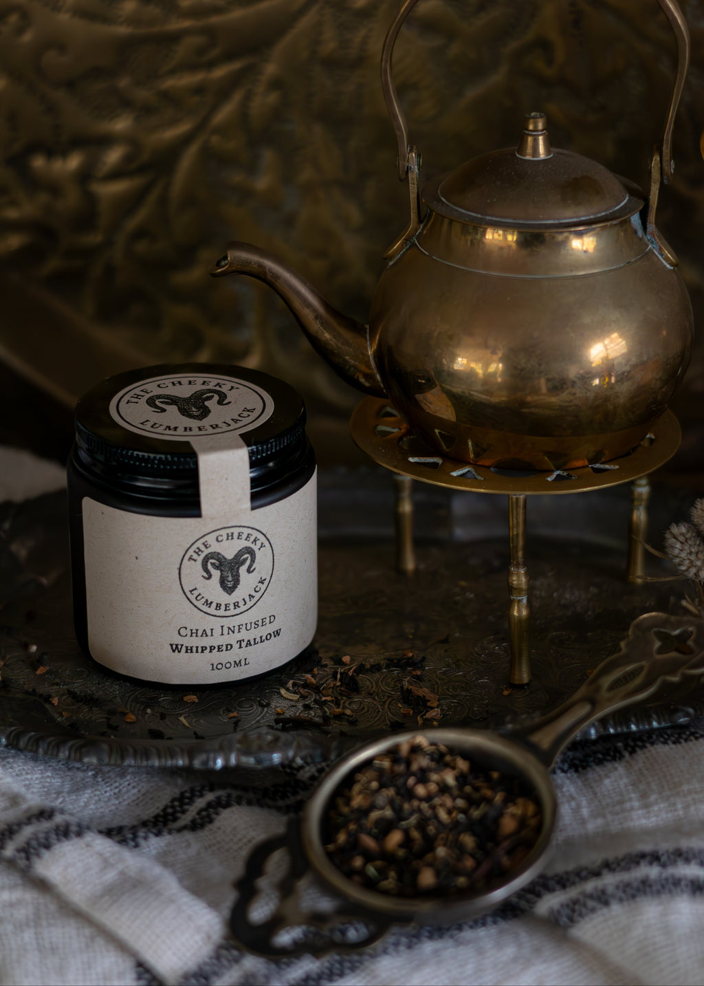 Jar of fern and sea chai infused whipped tallow with a brass teapot on a textured surface