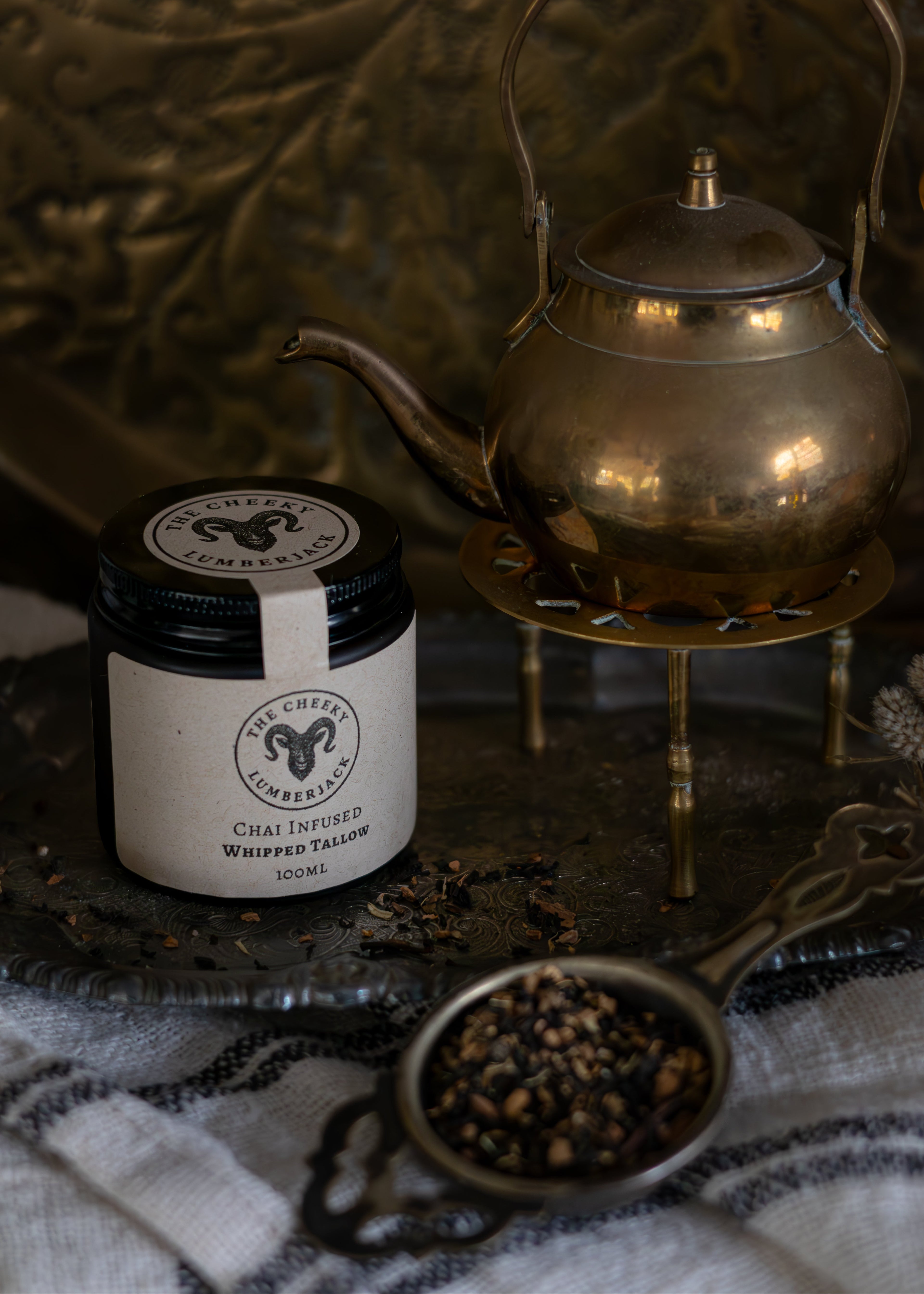 Jar of fern and sea chai infused whipped tallow with a brass teapot on a textured surface