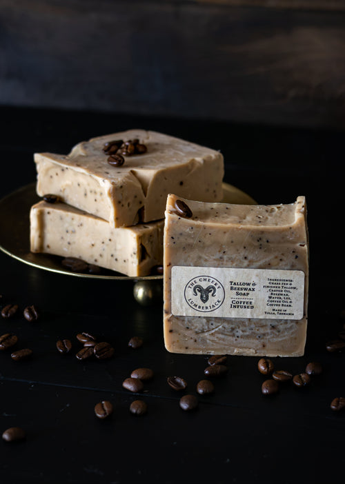 Three bars of Salome's Coffee & Vanilla Handmade Soap are displayed on a dark surface with coffee beans scattered around. One bar stands upright in the foreground showing a white label, while two others are stacked on a gold plate in the background.