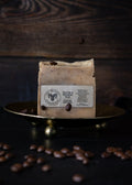 A rectangular bar of soap with a cream-colored label reading 'The Goat Herbs' sits on a small brass tray, with scattered coffee beans on a dark wooden surface and background.