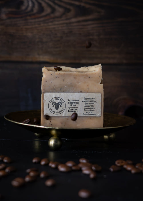 A rectangular bar of soap with a cream-colored label reading 'The Goat Herbs' sits on a small brass tray, with scattered coffee beans on a dark wooden surface and background.