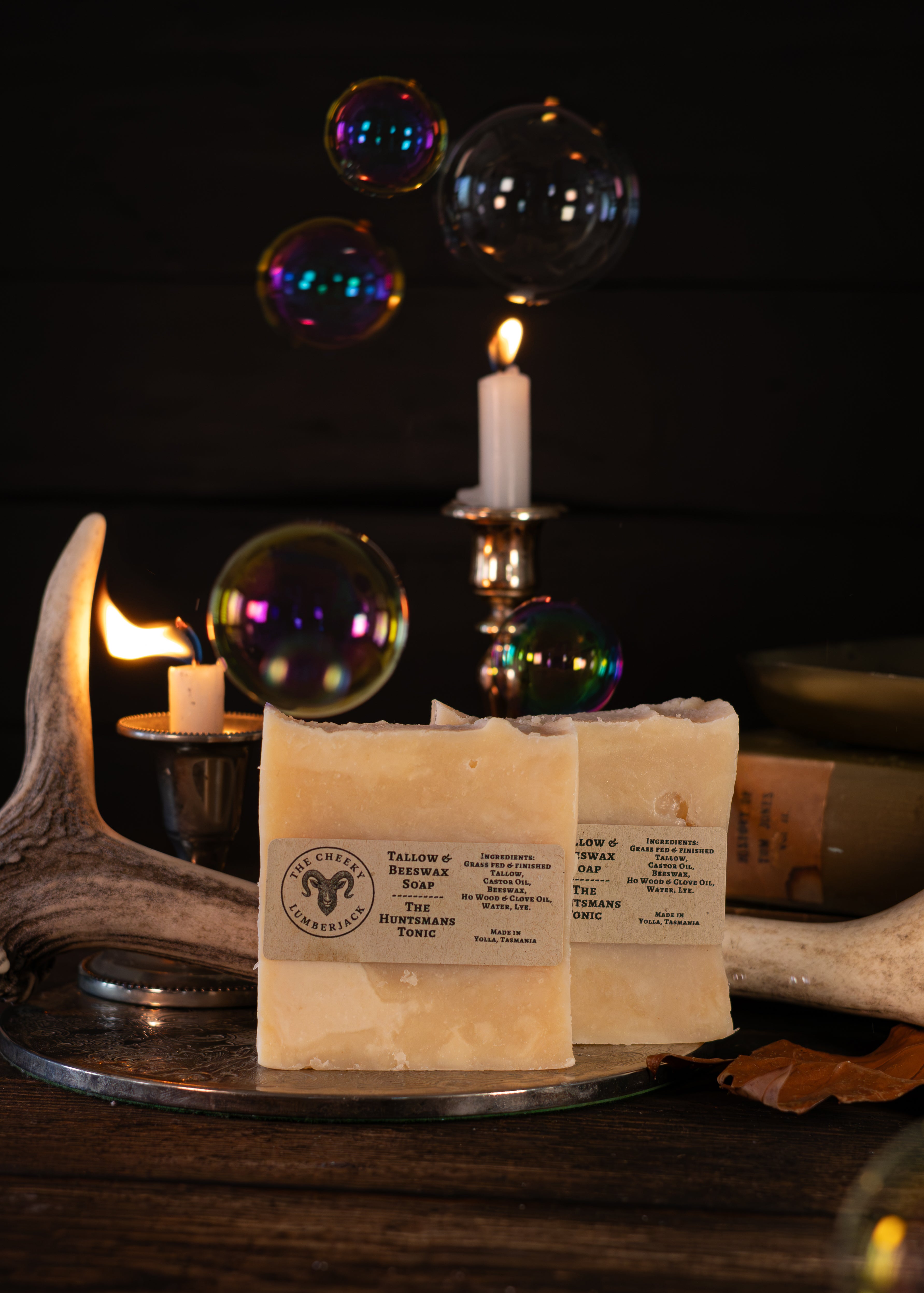 Two bars of handmade soap labeled 'Tallow & Beeswax Soap' by 'The Wildcrafted Shop' displayed on a rustic metal tray with candles, antlers, and floating bubbles against a dark background.