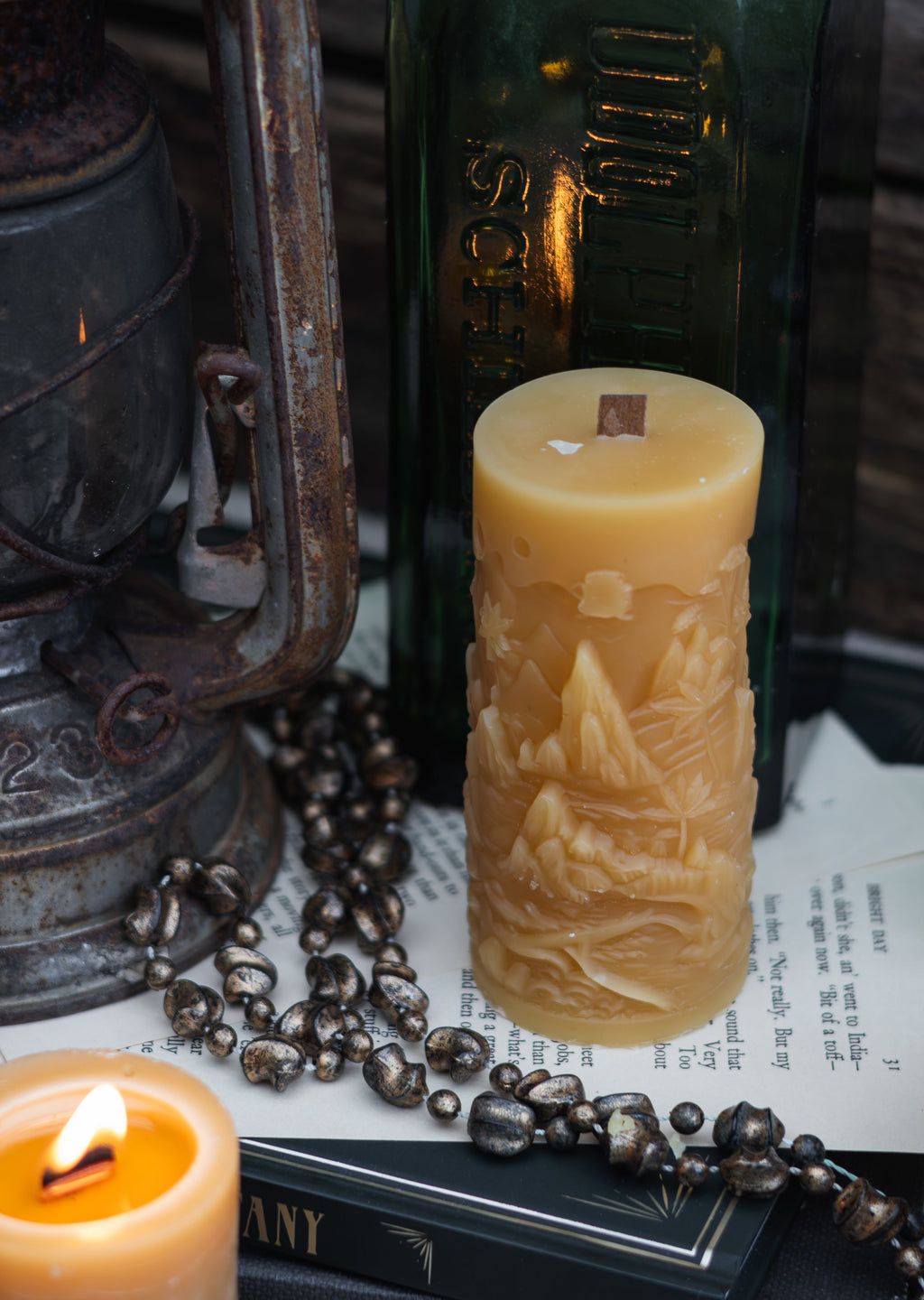 A carved beige pillar candle with a nautical mountain design stands on top of books, surrounded by a dark green glass bottle, a vintage lantern, a lit candle, and a beaded necklace.