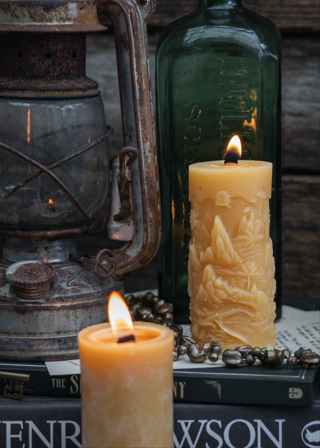 A styled tabletop scene with two lit cream-colored pillar candles, one cylindrical and one mountain landscape candle, alongside an antique lantern, a dark green glass bottle, books, and a beaded necklace against a rustic wooden background.