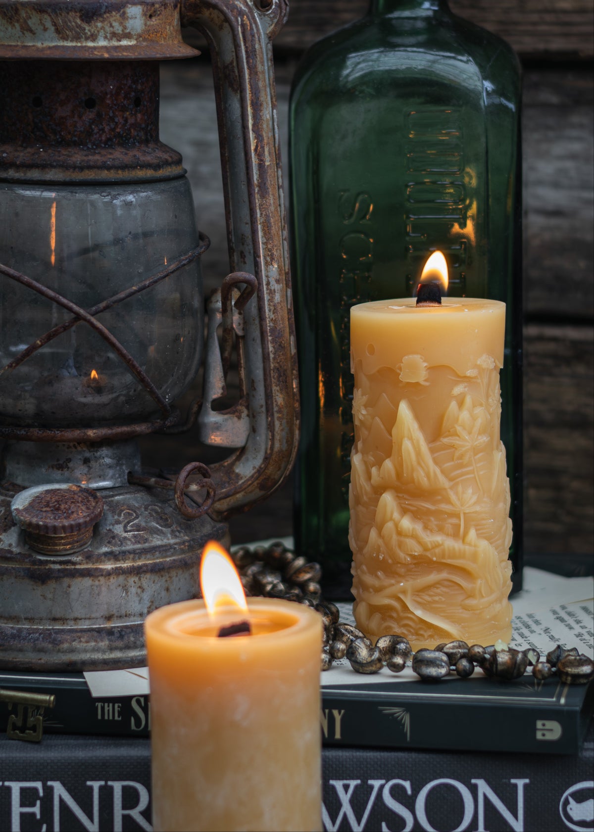A styled tabletop scene with two lit cream-colored pillar candles, one cylindrical and one mountain landscape candle, alongside an antique lantern, a dark green glass bottle, books, and a beaded necklace against a rustic wooden background.