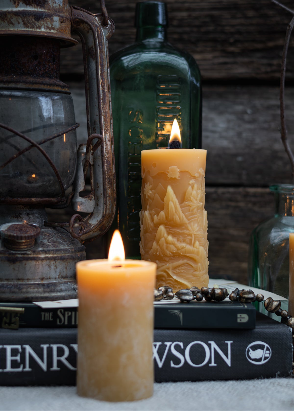 Two lit beeswax pillar candles, one short and one tall with a mountain relief design, styled on books with a dark green glass bottle and an antique lantern in the background.