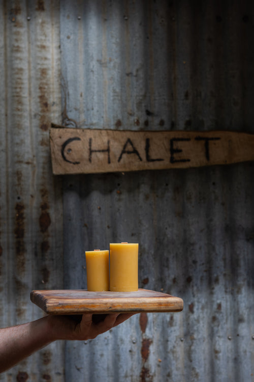Two yellow candles on a wooden board held by a hand against a corrugated metal wall with 'CHALET' sign.