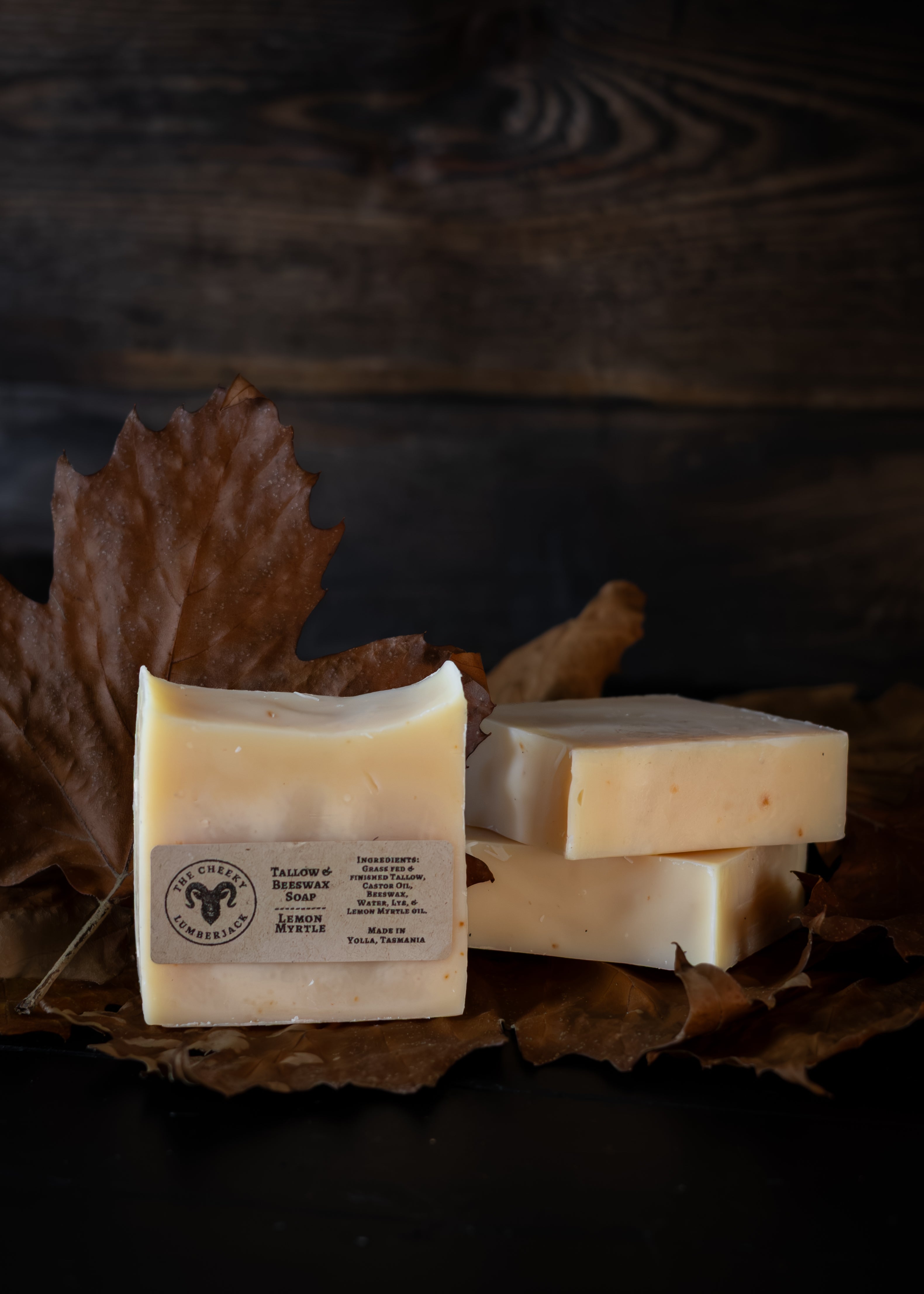 Three rectangular bars of handmade soap in warm cream and pale yellow tones, arranged on dried brown leaves against a dark rustic wooden background. One bar in the foreground has a white paper label with a circular logo and text.