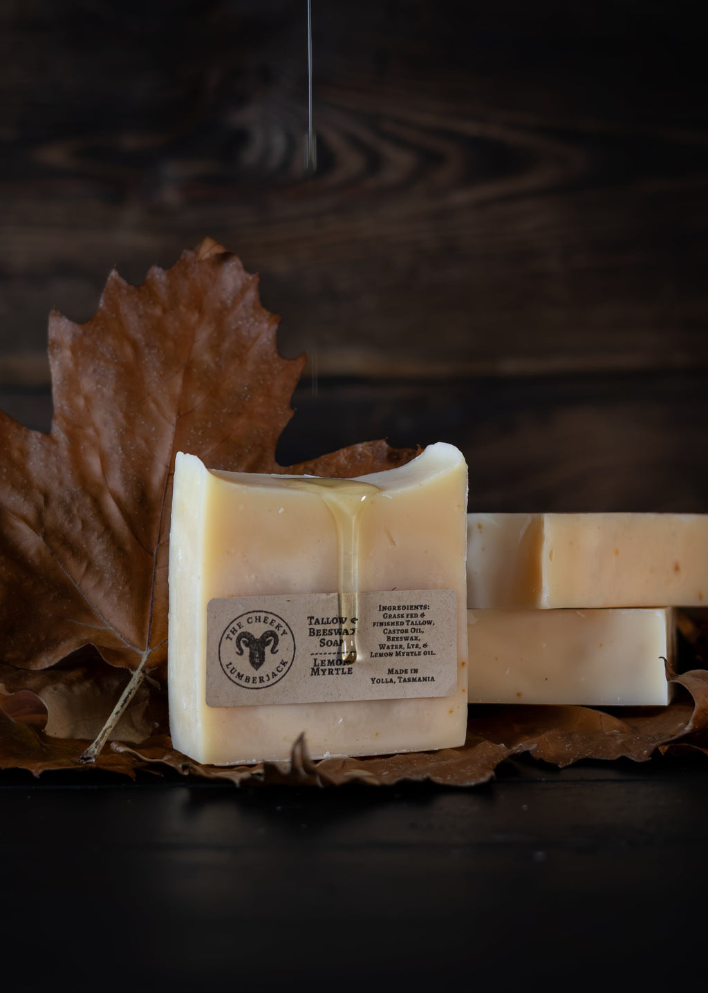 Three bars of handmade soap in a warm cream color, with one bar in the foreground showing a label and a glossy drizzle of honey, styled on brown autumn leaves against a dark wood background.