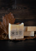 Three bars of handmade soap in a warm cream color, with one bar in the foreground showing a label and a glossy drizzle of honey, styled on brown autumn leaves against a dark wood background.