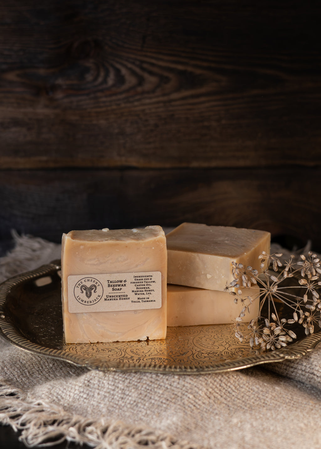 Two rectangular bars of soap in a warm pale yellow tone, one standing upright with a paper label and one stacked behind it, displayed on a decorative gold-toned tray with dried flowers against a dark rustic wood background.