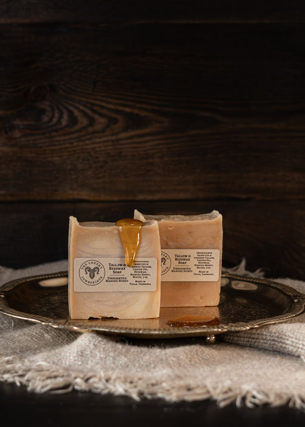 Two rectangular bars of soap labeled 'Tasmanian Honey Bar' resting on a silver tray with a piece of dripping honey on the front bar, set against a dark wood background.