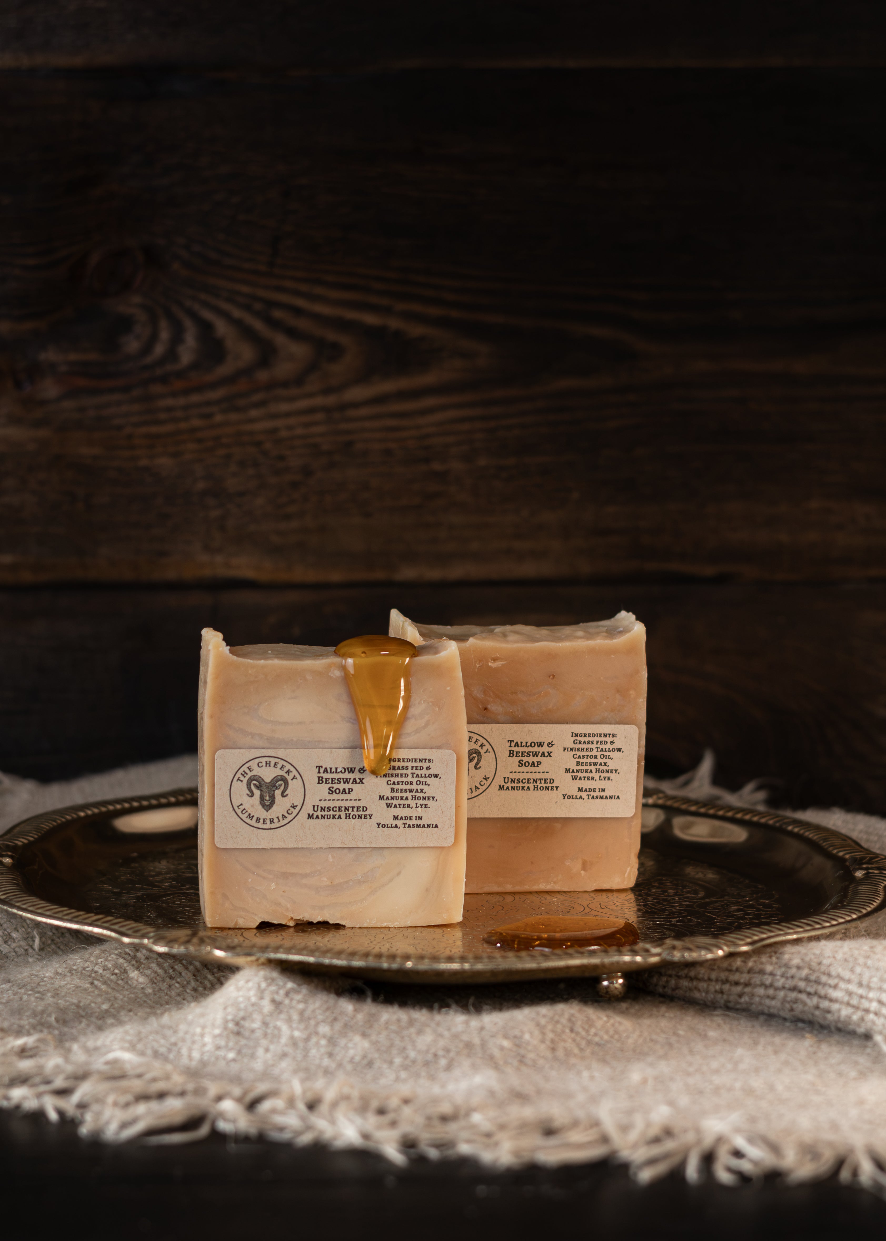 Two rectangular bars of soap labeled 'Tasmanian Honey Bar' resting on a silver tray with a piece of dripping honey on the front bar, set against a dark wood background.