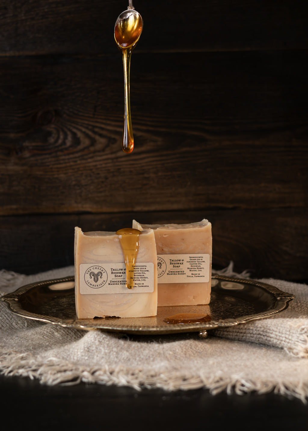 Two rectangular bars of soap in cream-colored paper packaging sit on a round silver tray, with golden honey dripping from the top and a honeycomb pattern printed on the front label. The tray is placed on a textured white cloth against a dark wood background.