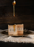 Two rectangular bars of soap in cream-colored paper packaging sit on a round silver tray, with golden honey dripping from the top and a honeycomb pattern printed on the front label. The tray is placed on a textured white cloth against a dark wood background.