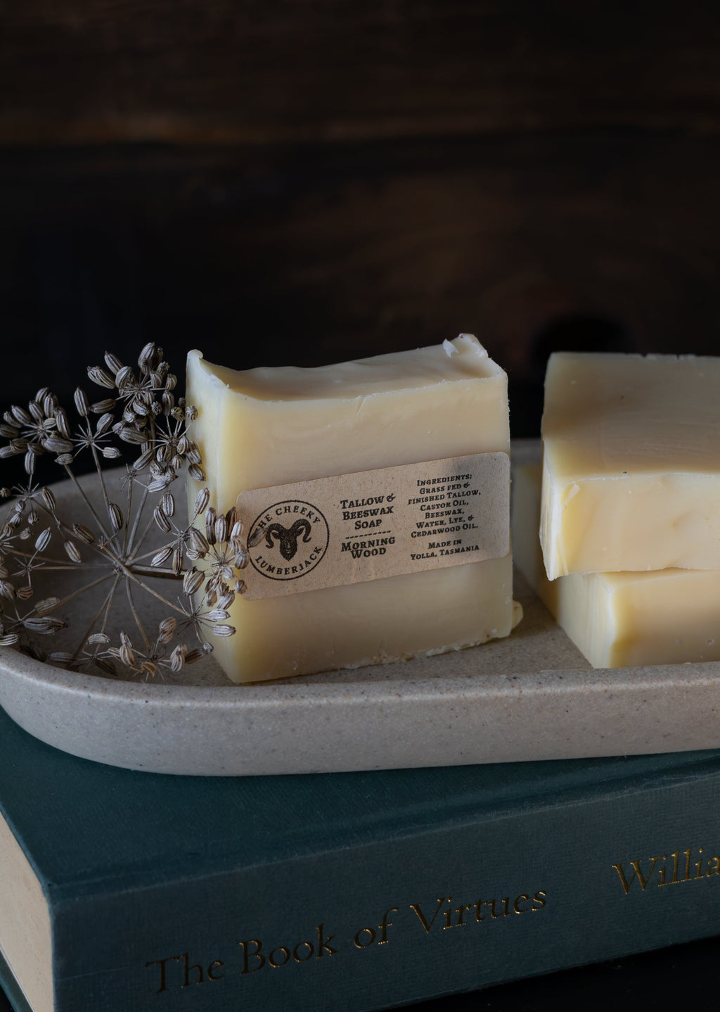 Two bars of handmade tallow castor oil beeswax soap in off-white cream, one wrapped with a paper label and one unwrapped, displayed on a speckled ceramic dish with dried flowers on top of a teal book against a dark wood background.