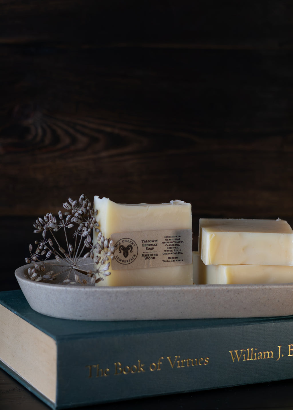 Three bars of pale yellow handmade soap arranged on a gray ceramic tray, with dried flowers beside one bar and a teal book underneath, set against a dark wood background.