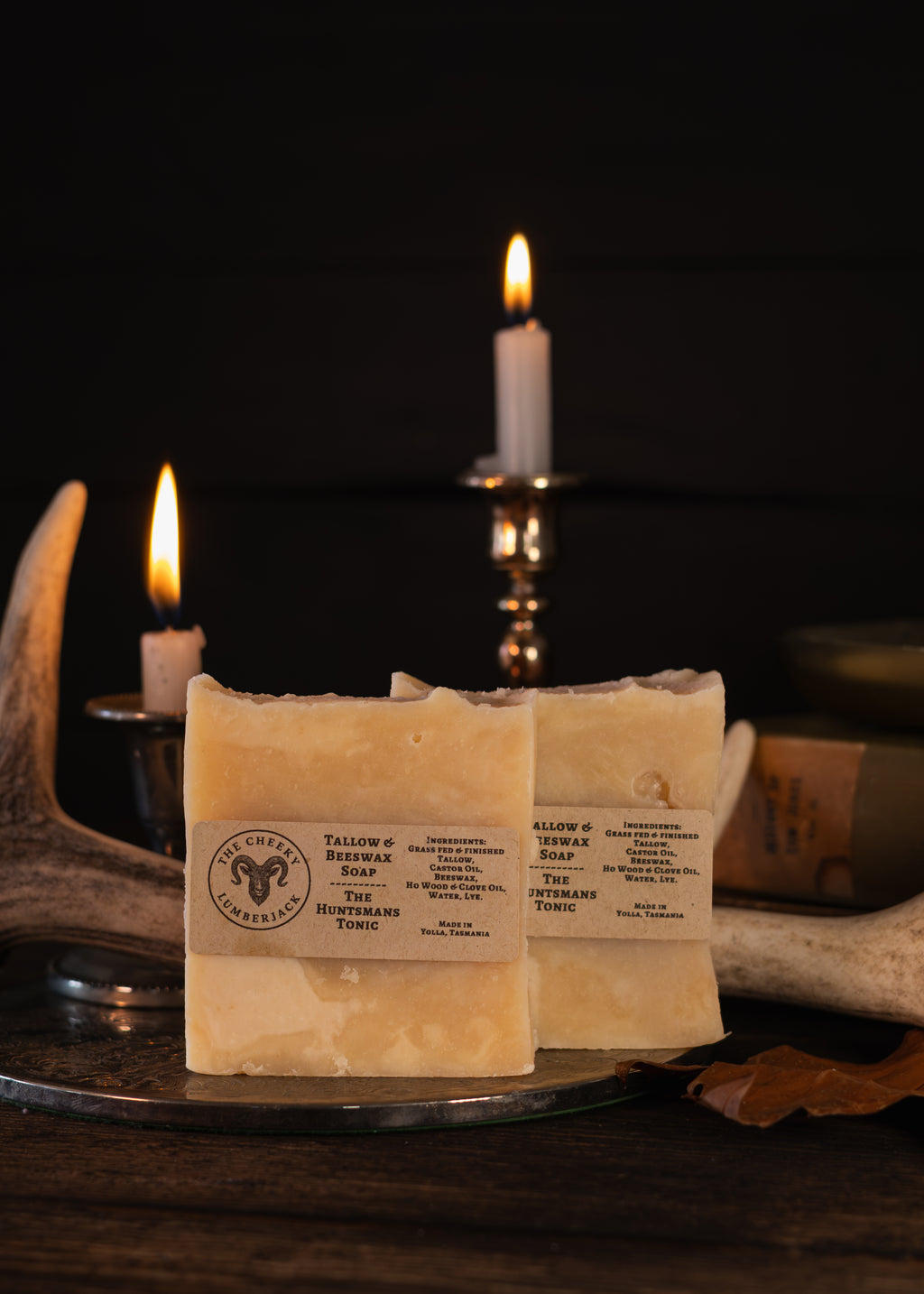 Two bars of handmade soap labeled 'The Huntsman's Tonic' resting on a metal tray, styled with lit candles, antlers, and a dark rustic background.